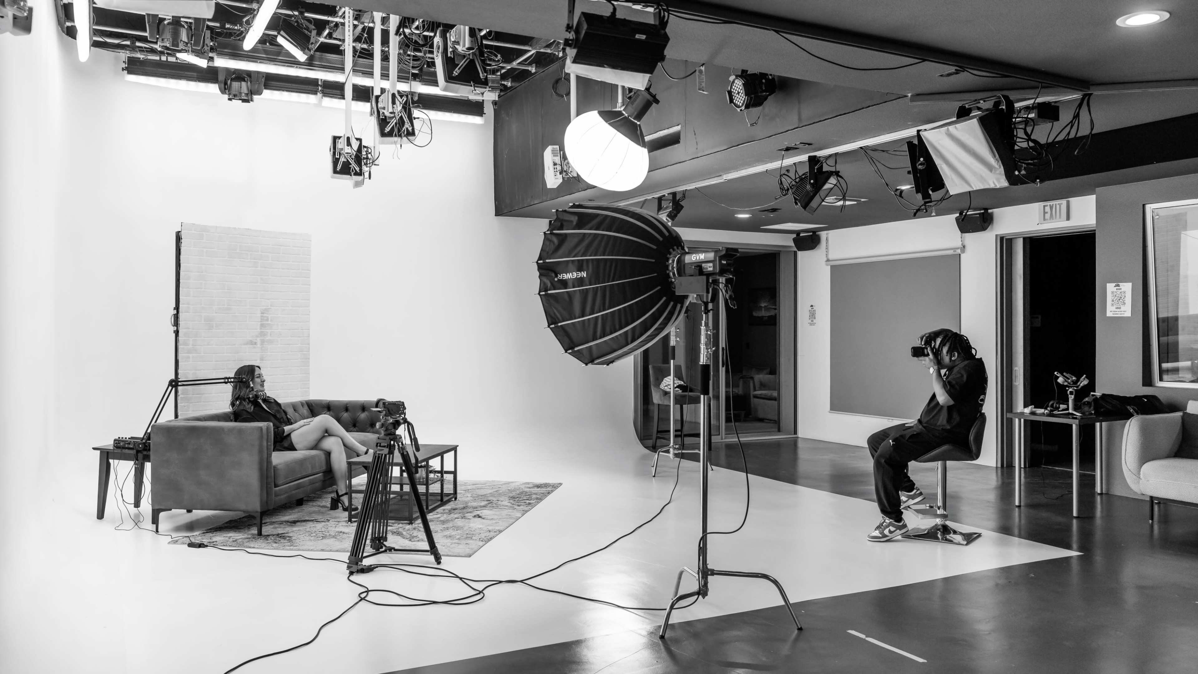 A photographer is seated on a stool, capturing an image of a model posed on a couch in a well-lit studio setting.