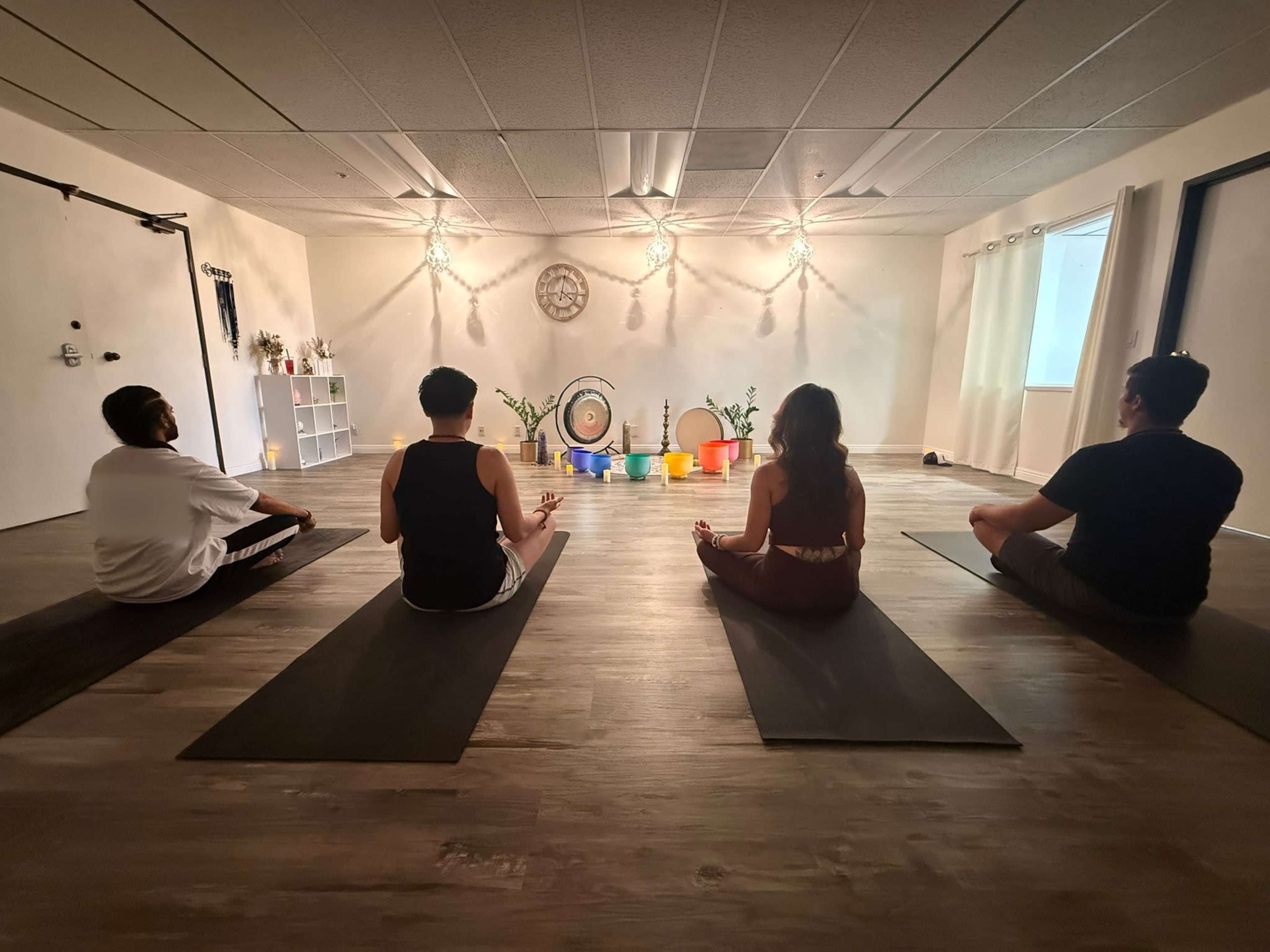 Four individuals sit in a yoga studio on mats facing a wall adorned with plants and a decorative clock under soft lighting.
