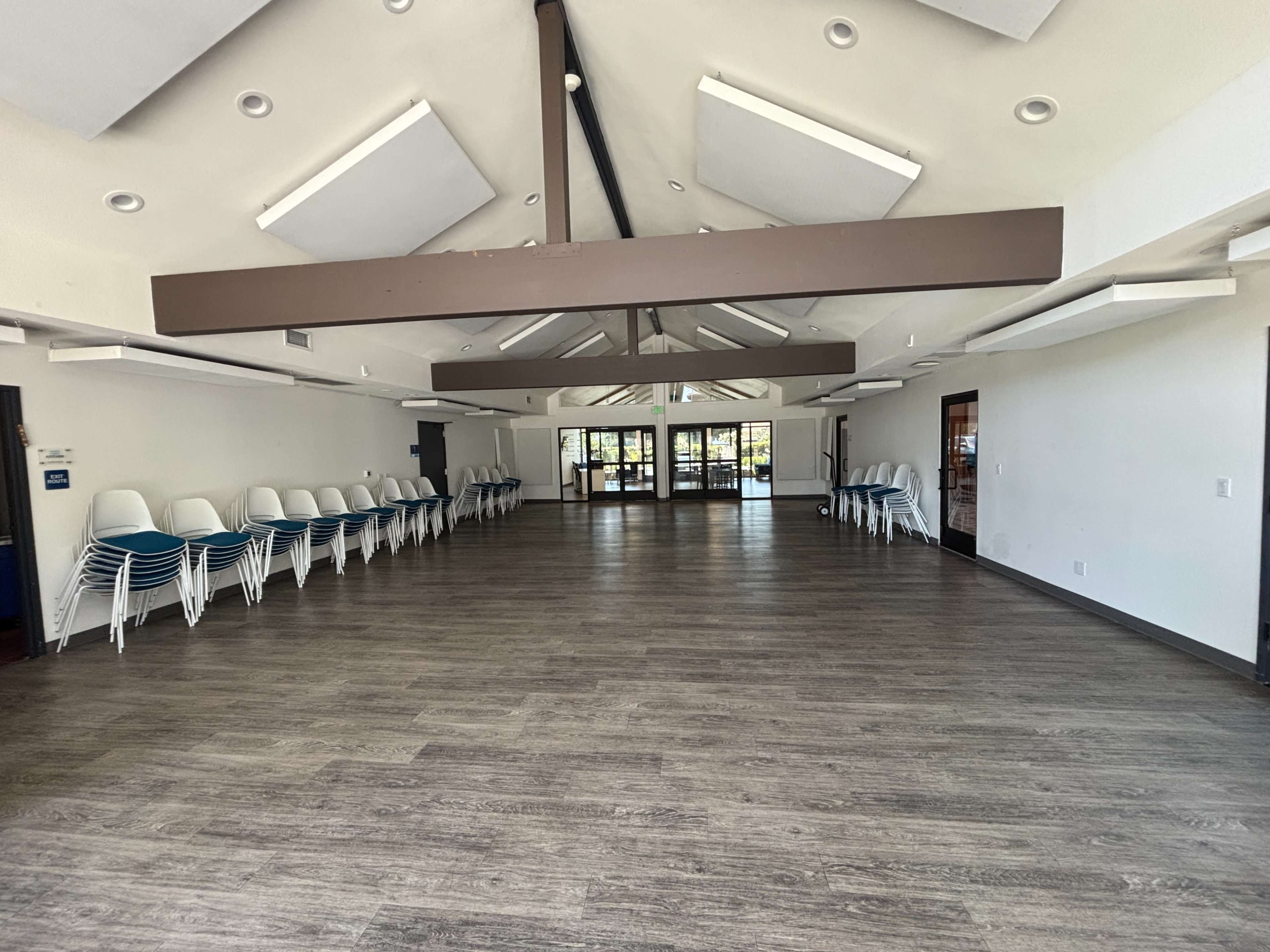 The image shows an empty interior space with wooden flooring, a high ceiling, and stacks of white chairs along one wall.