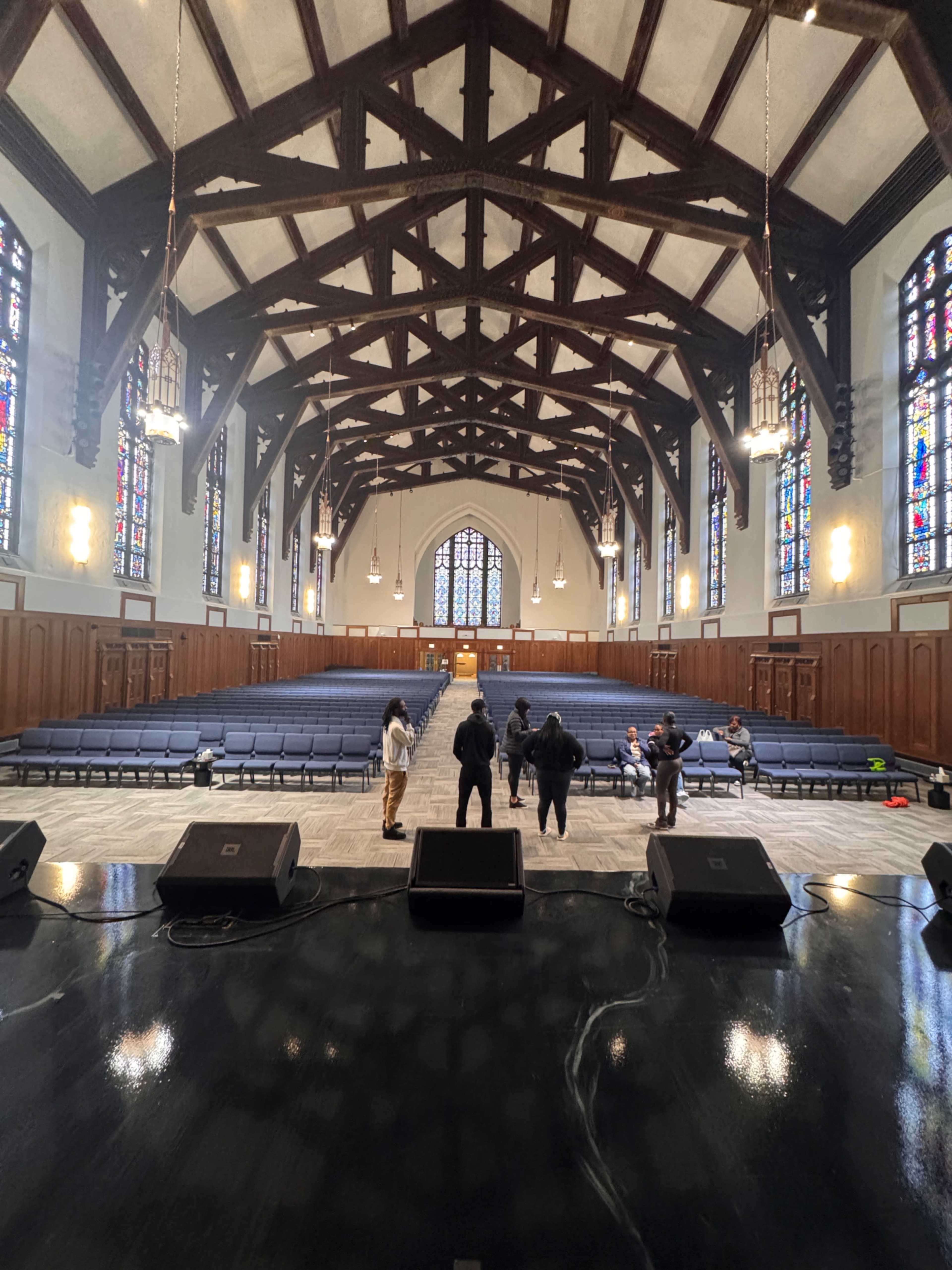 The image shows a large indoor auditorium with a wooden ceiling, stained glass windows, and rows of chairs facing a stage where several individuals are gathered.