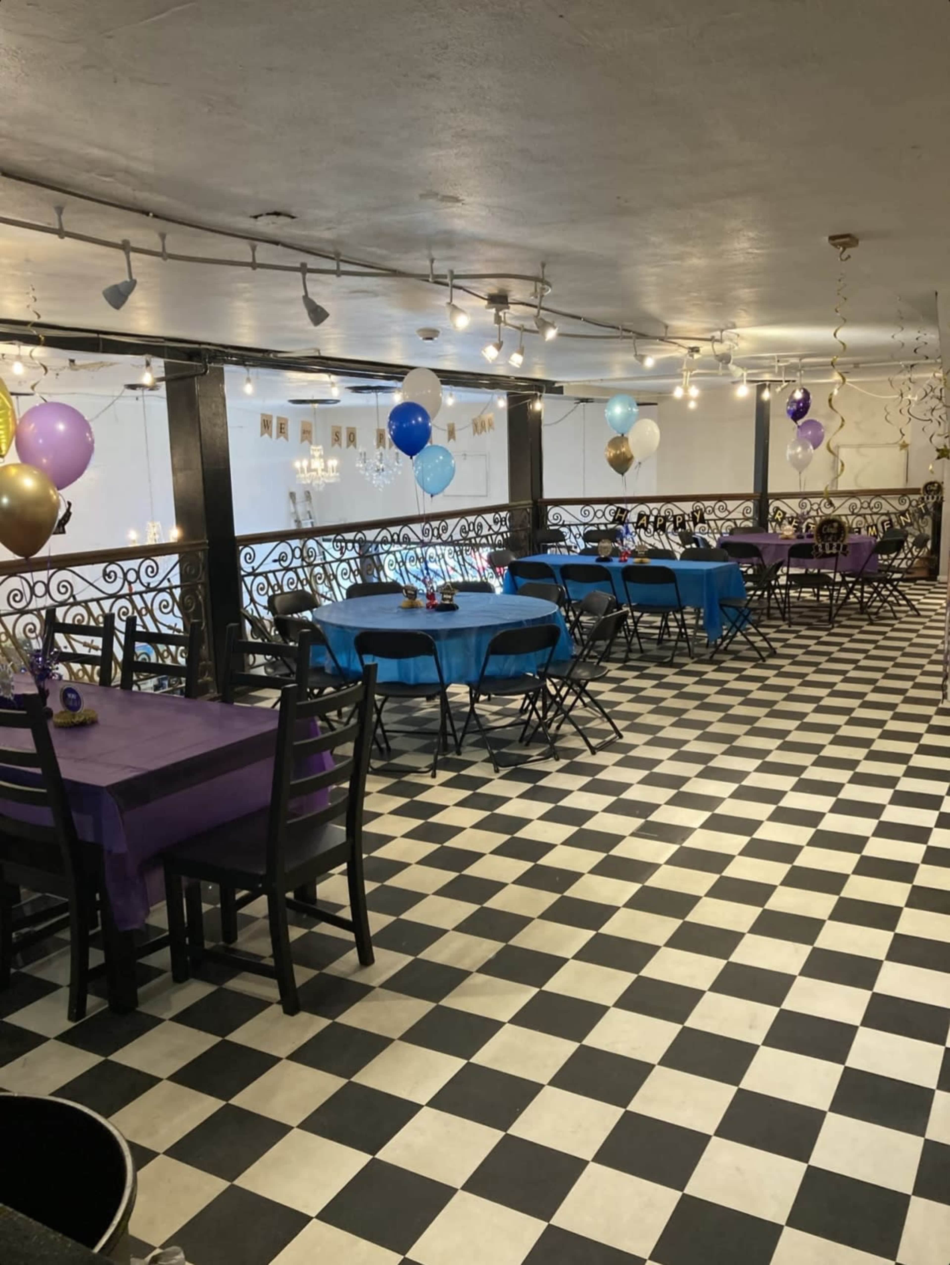 An indoor venue with black and white checkered flooring, featuring tables covered in purple and blue cloths, and balloons in various colors decorations.