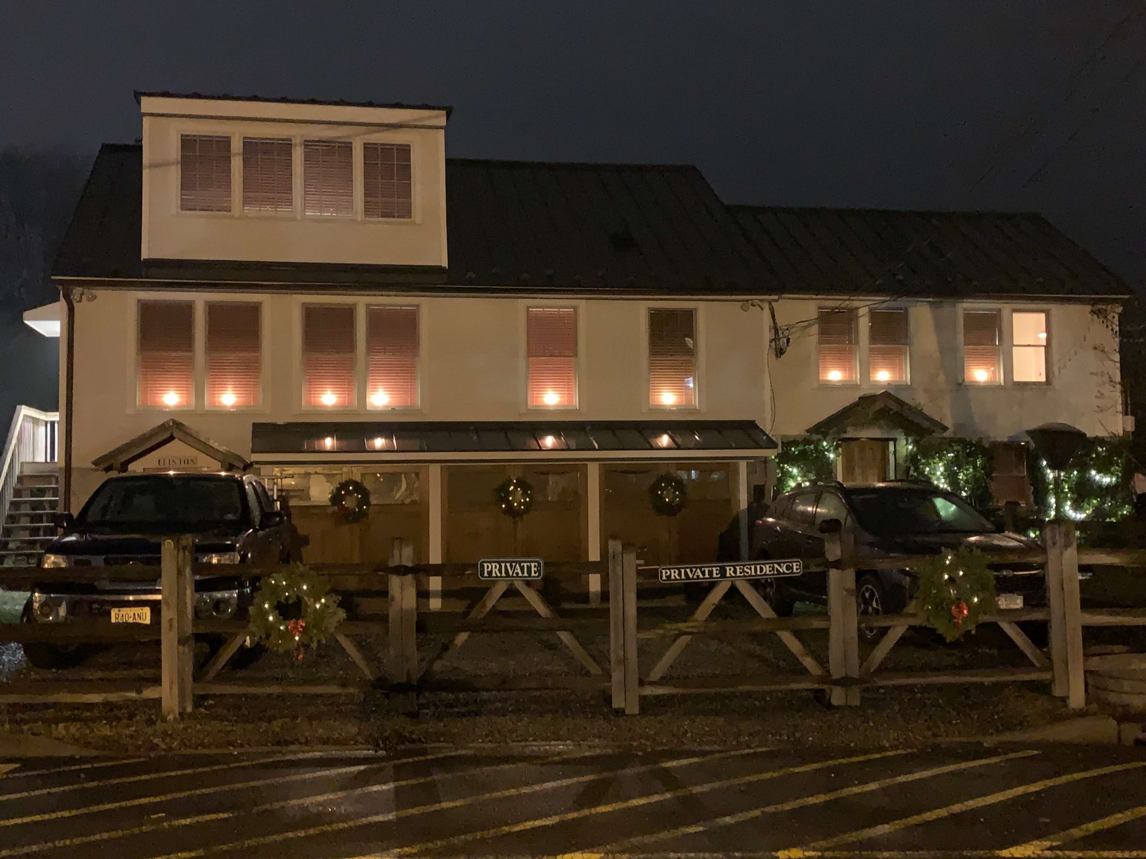 A well-lit private residence features decorative wreaths on the front and two parked vehicles in front of a fence at night.