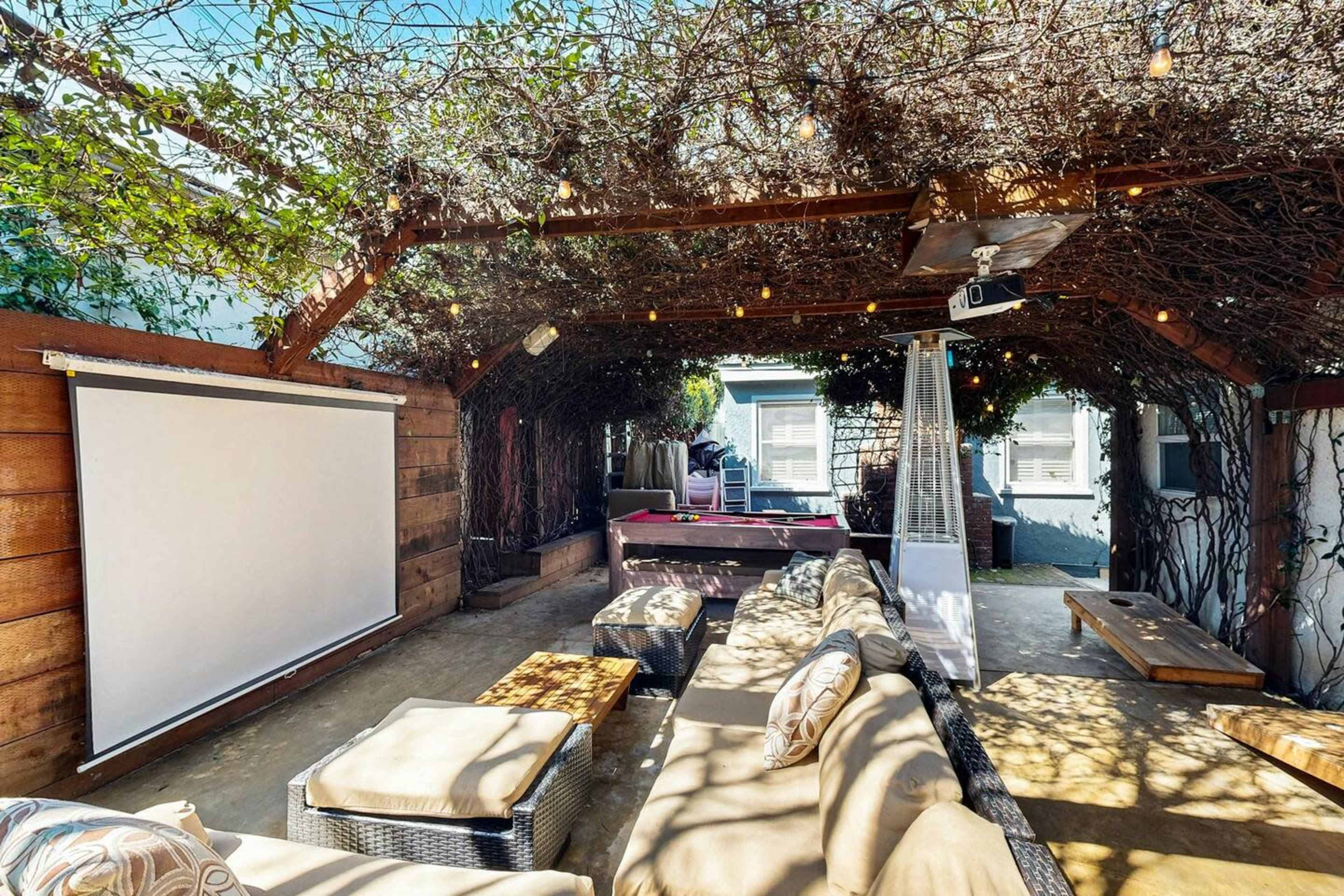 A shaded outdoor lounge area with cozy seating, a large projector screen, and twinkling lights draped above.