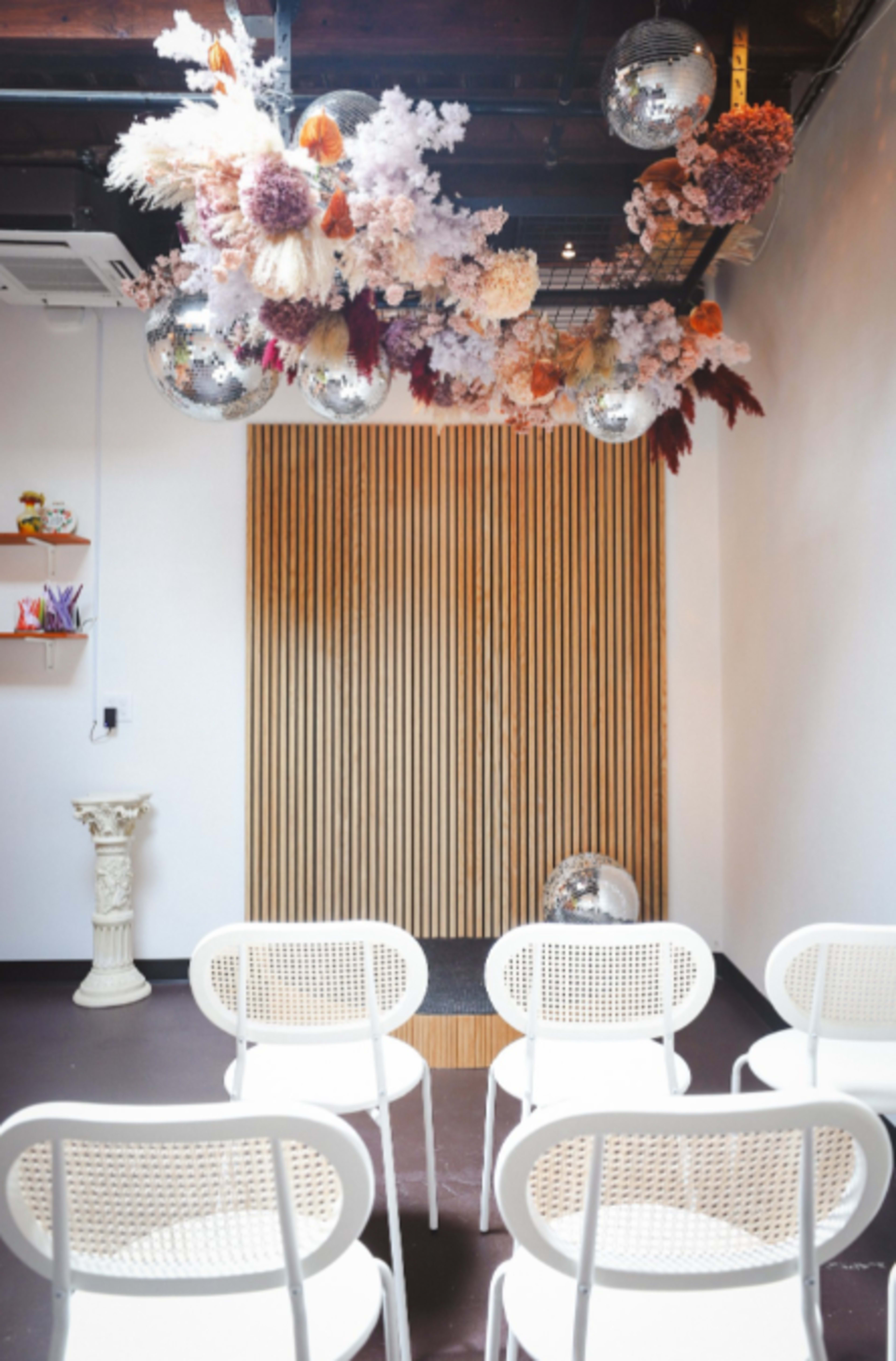 A minimalist event space featuring white chairs arranged in front of a wooden wall, adorned with a floral installation and disco balls overhead.