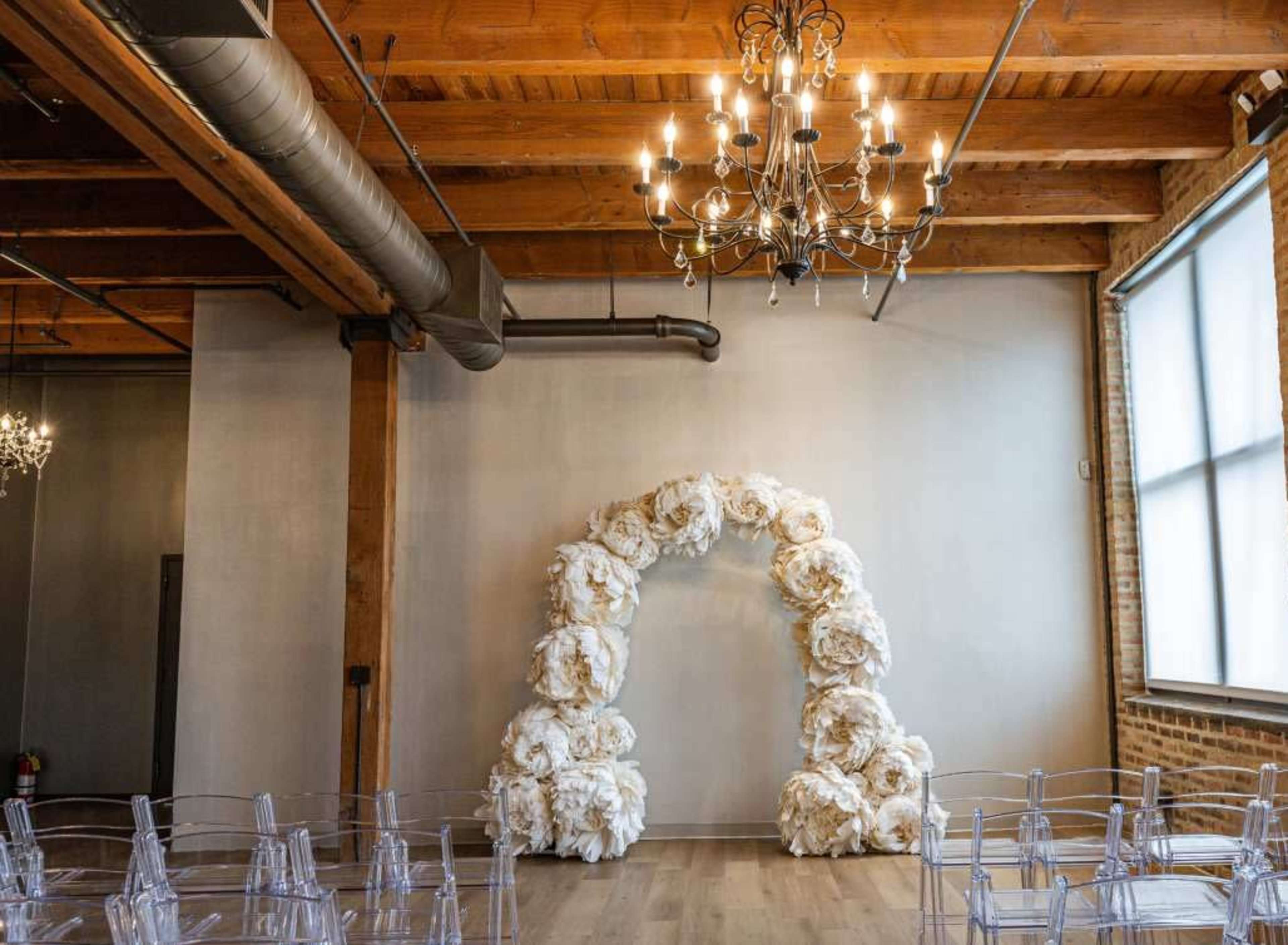 The image shows a spacious event space with wooden beams and a floral arch at the front, surrounded by transparent chairs arranged in rows.