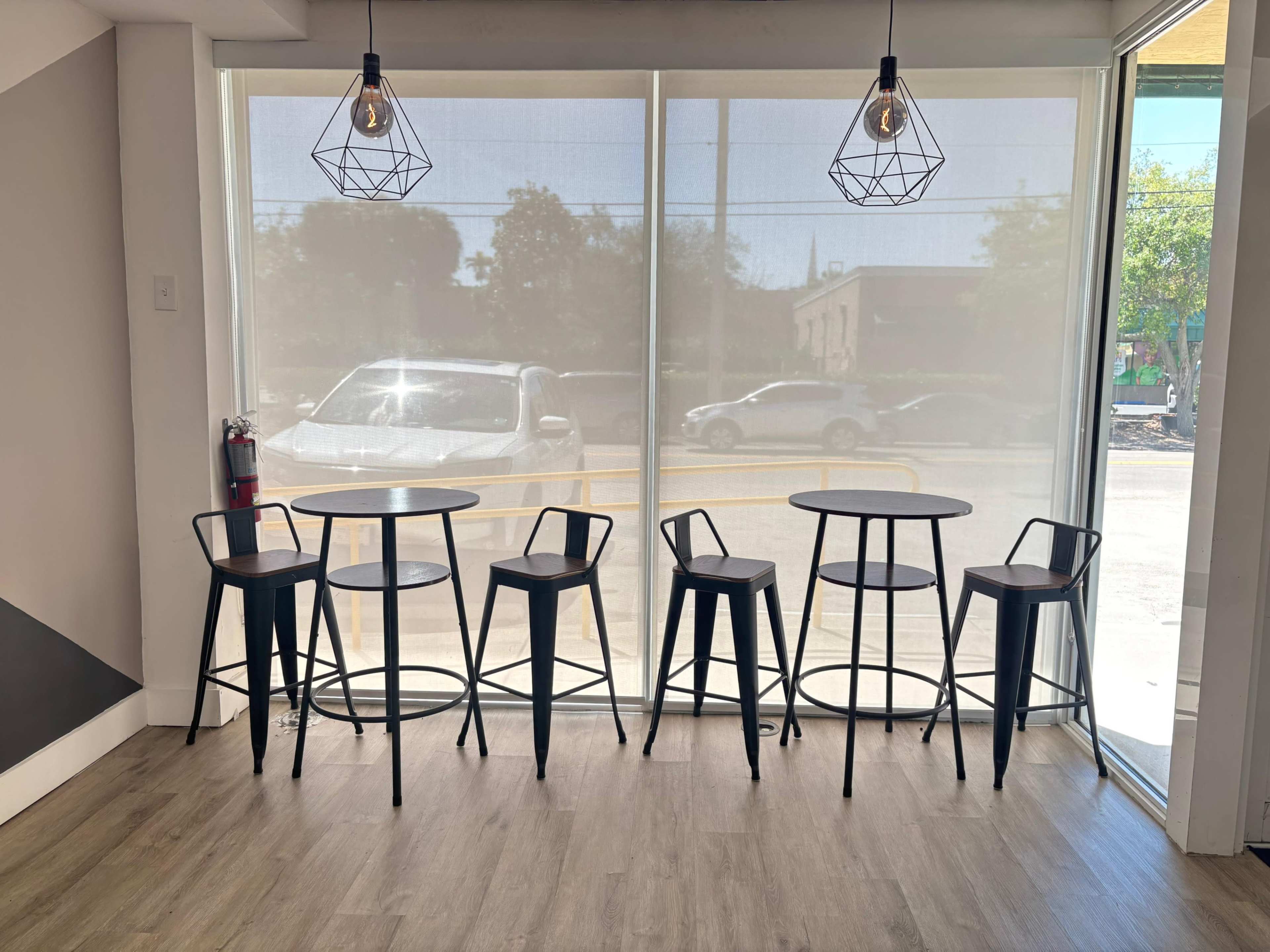 The image features a row of black metal high tables and chairs placed by a large window with natural light streaming in.