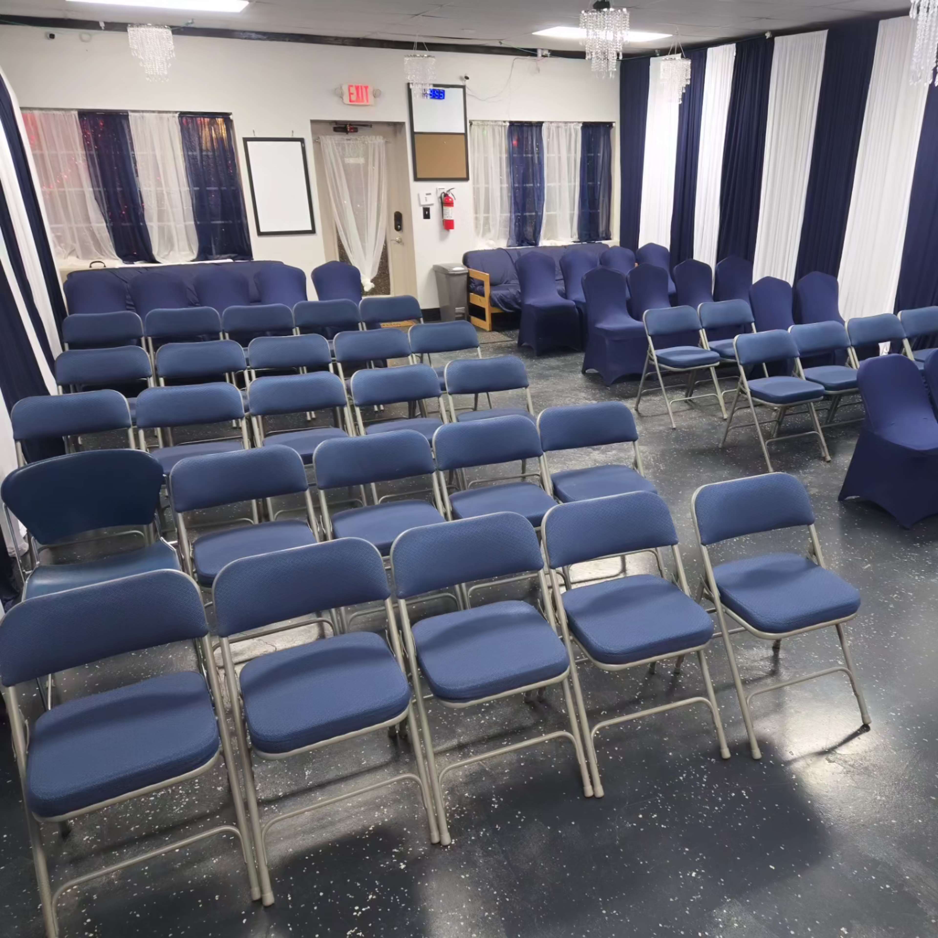 The image shows a seminar room set up with rows of blue chairs facing a front area, with white and navy drapes decorating the walls.