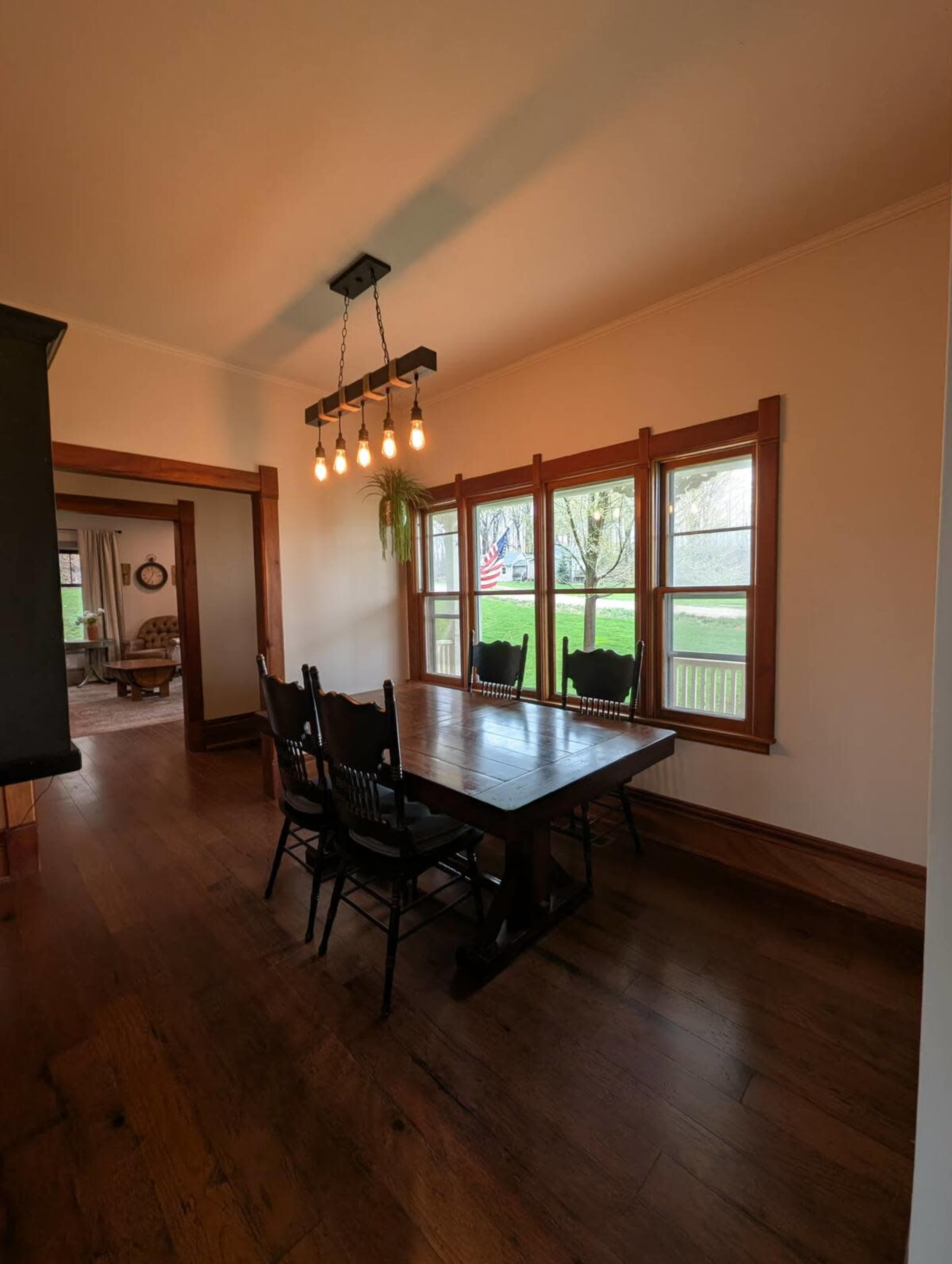A spacious dining room features a wooden table surrounded by black chairs, illuminated by a hanging light fixture above, with large windows showcasing a view of greenery outside.