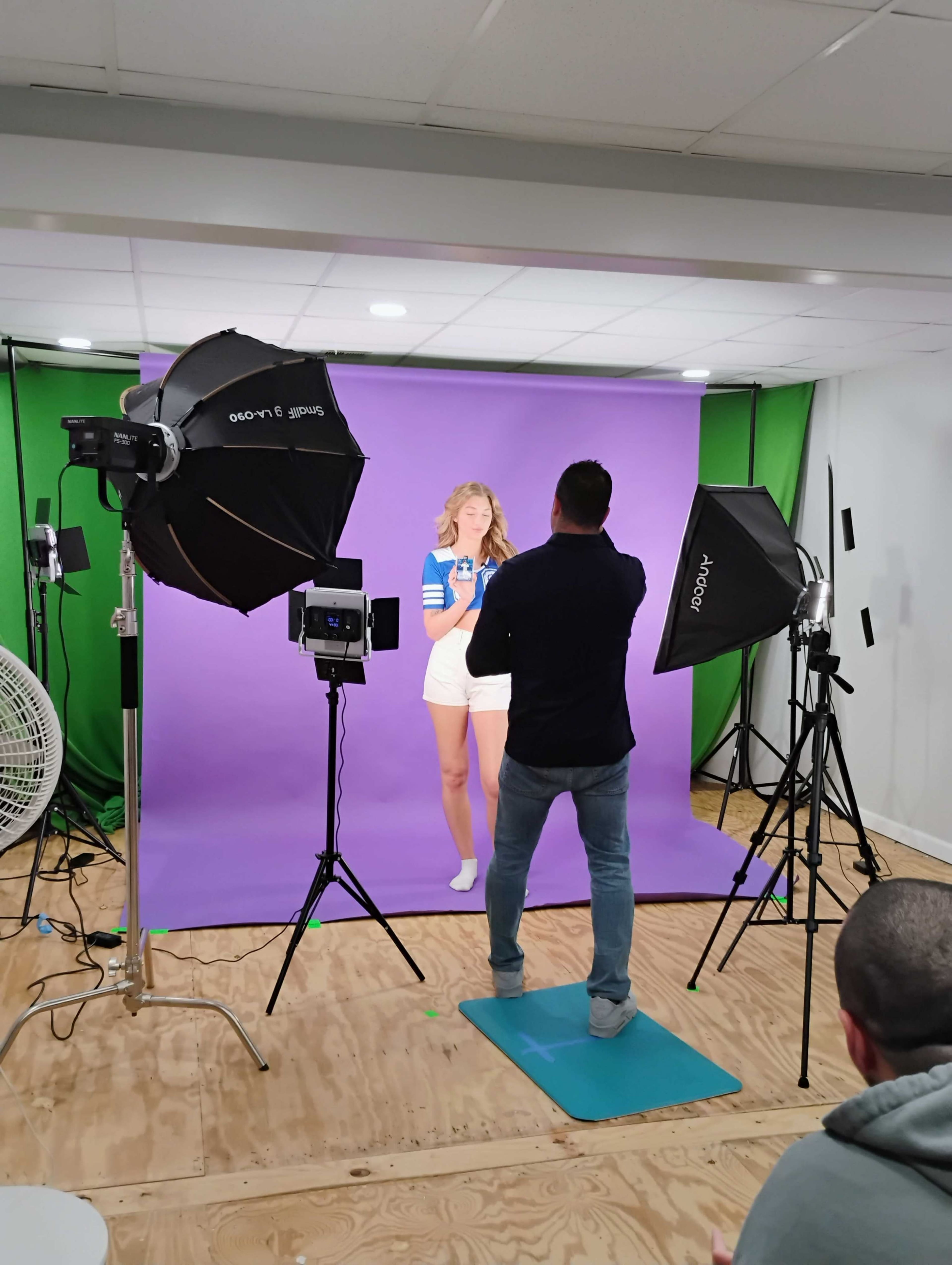 A model poses against a purple backdrop while a photographer adjusts lighting equipment in a studio setting.