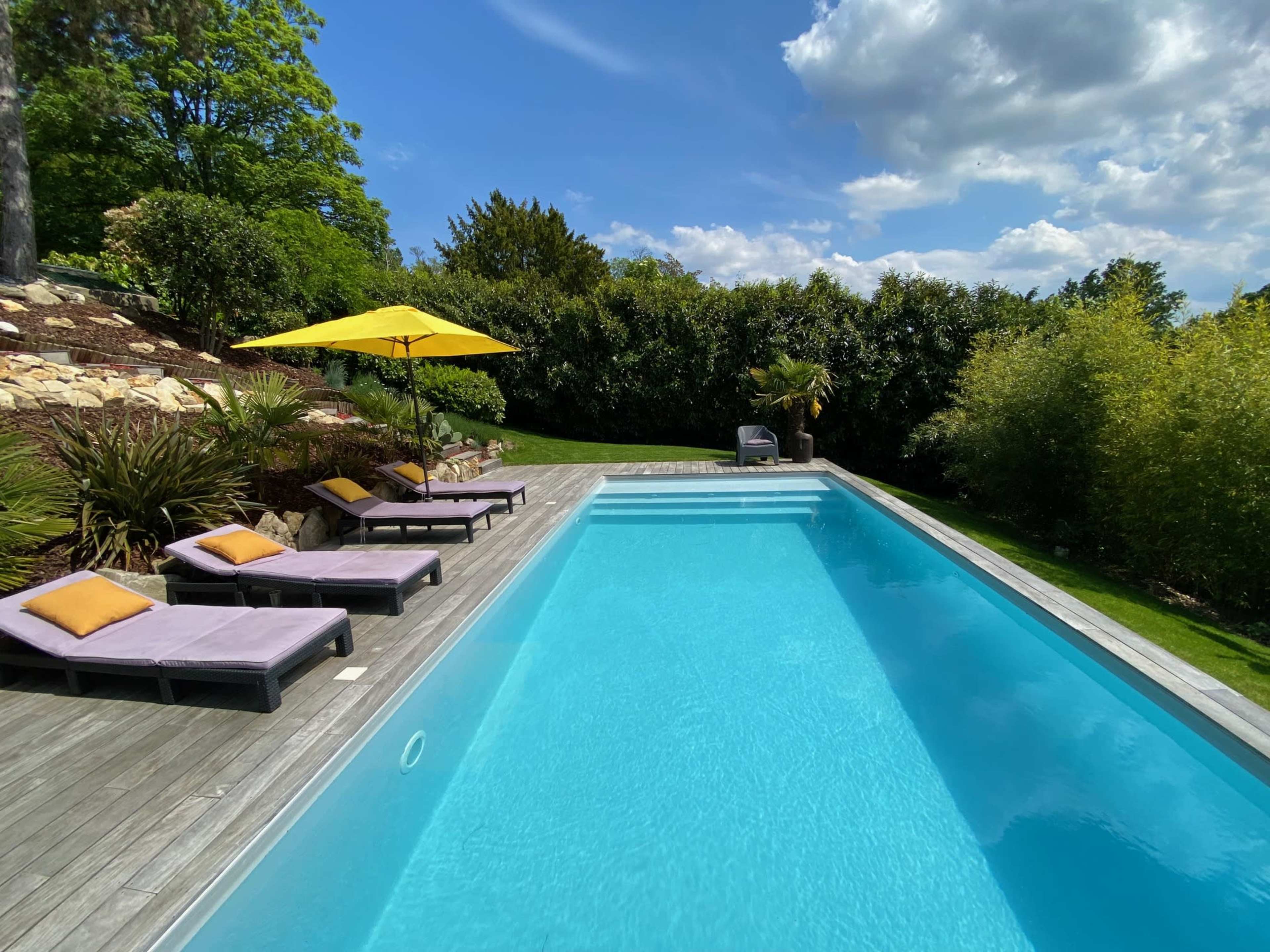A rectangular swimming pool is surrounded by wooden decking and lounge chairs with cushions, with an umbrella providing shade nearby and lush greenery in the background.