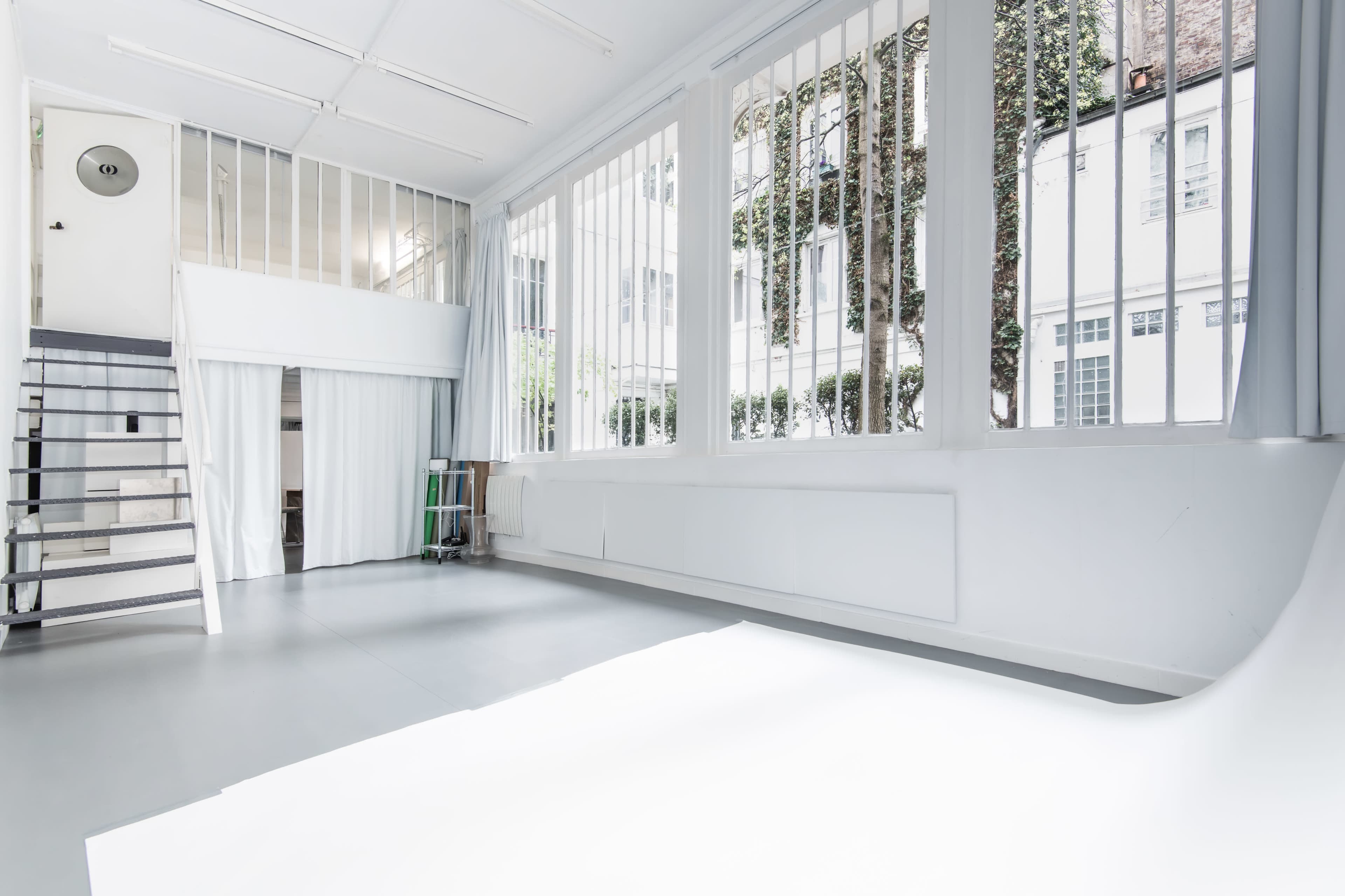 A spacious, minimalist studio space features large windows with vertical bars and a white floor, designed for natural light.