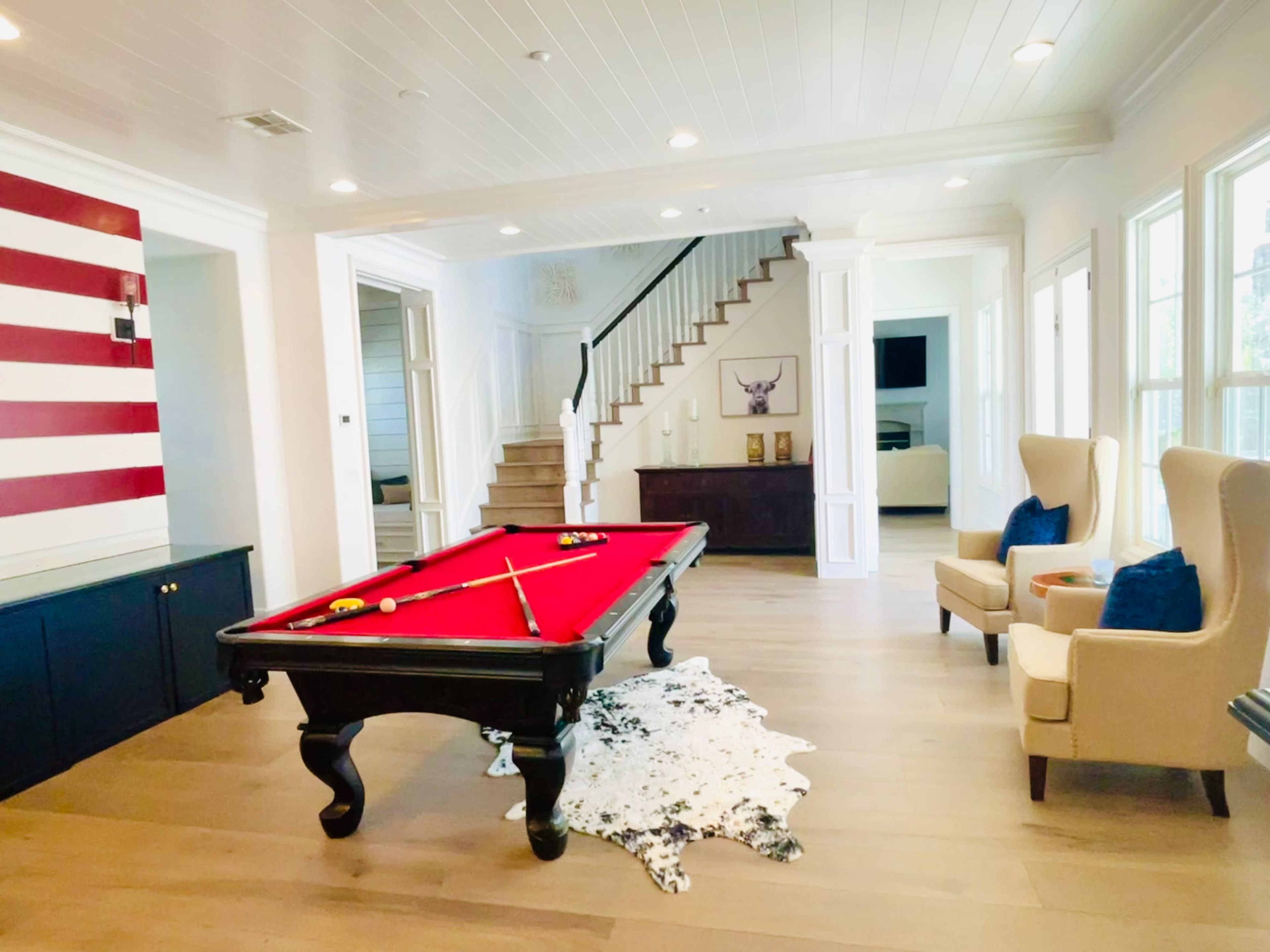 The room features a red felt pool table, a decorative cowhide rug, and a staircase leading to an upper level, complemented by large windows allowing natural light.