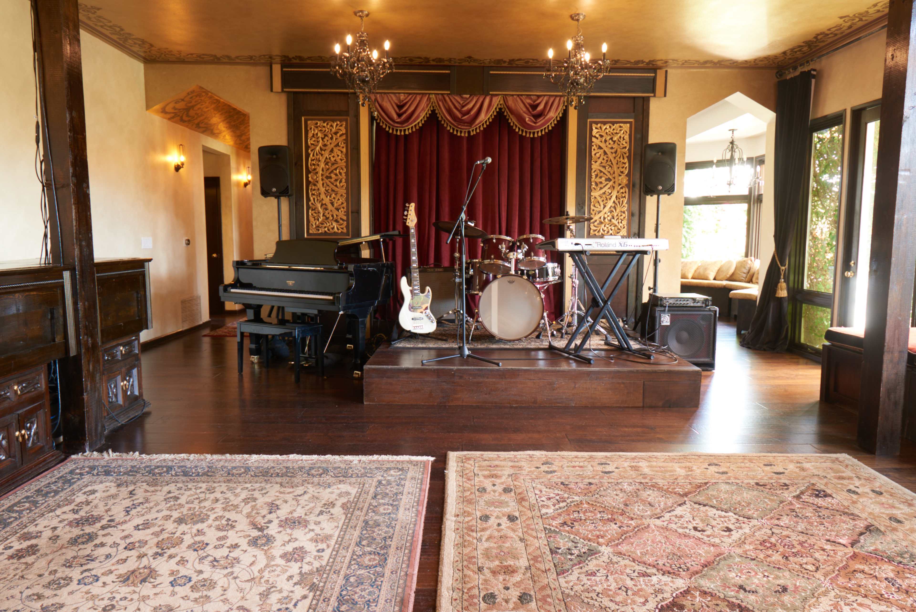 A stage setup for a performance, featuring a grand piano, a drum kit, electric guitar, and keyboard, with decorative curtains and a cozy seating area in the background.