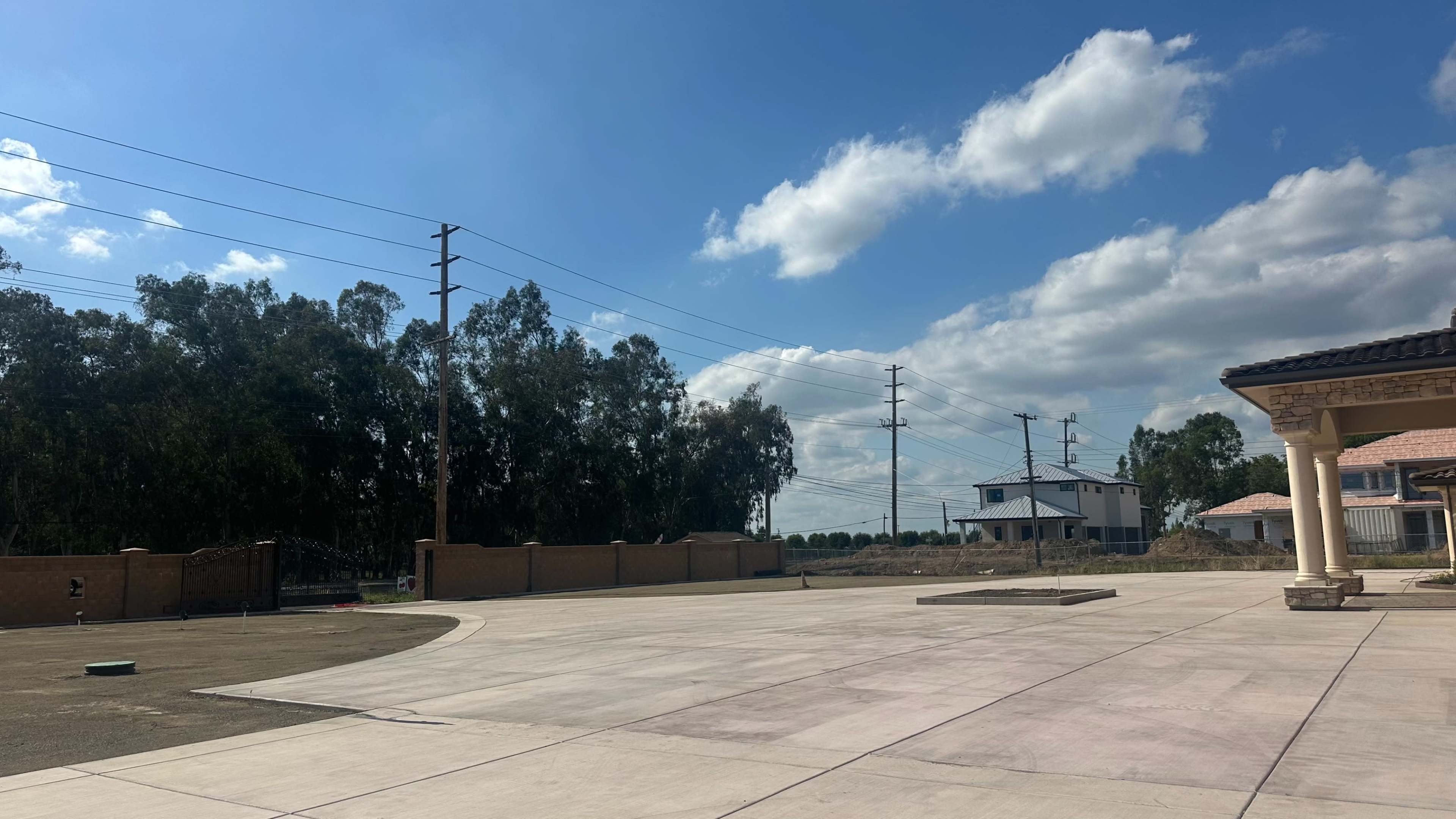 The image shows a wide, open area with a concrete surface, flanked by trees and power lines under a partly cloudy sky.