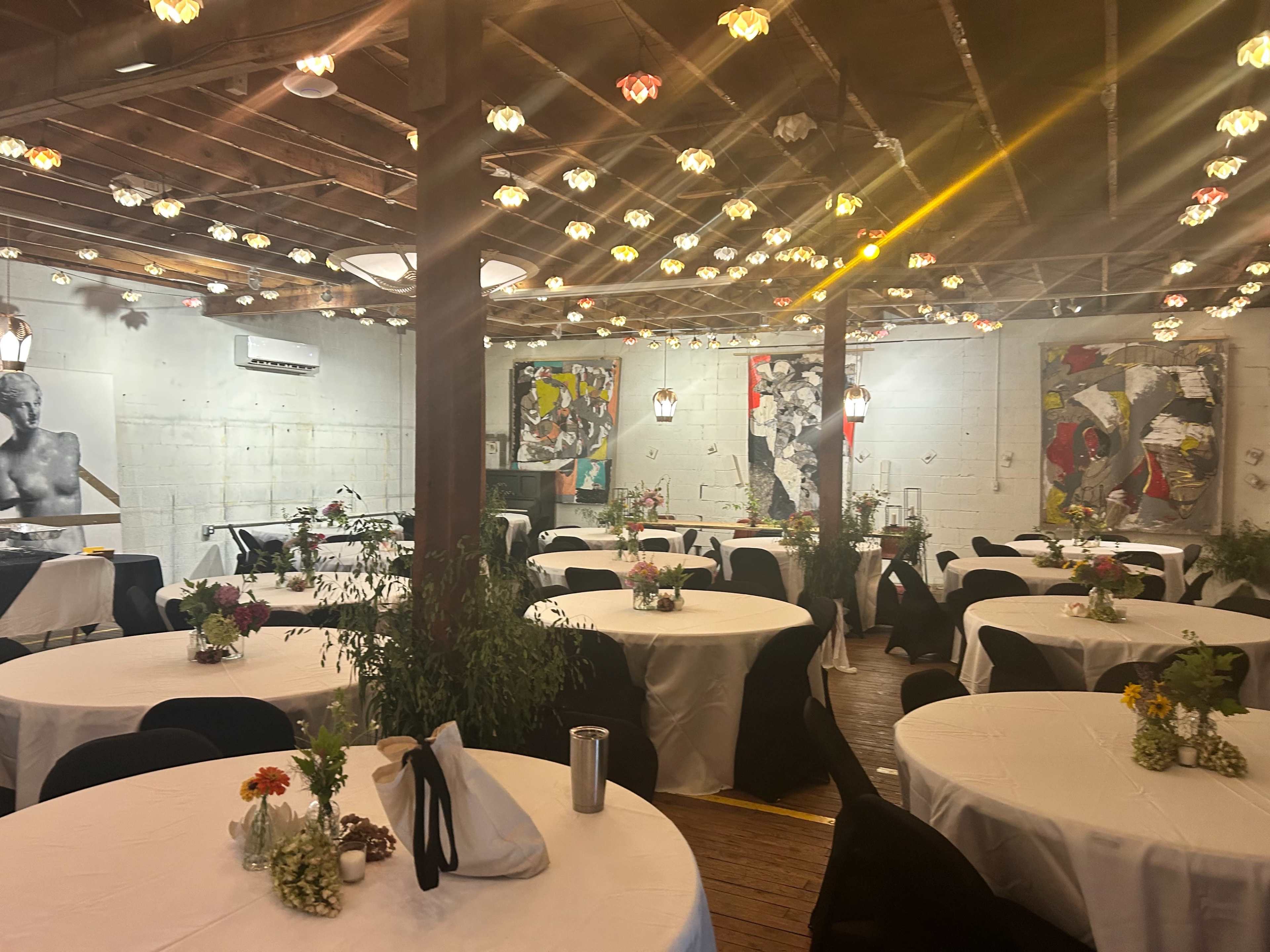 The image shows a decorated event space with round tables covered in white tablecloths, adorned with floral centerpieces and illuminated by hanging lights.