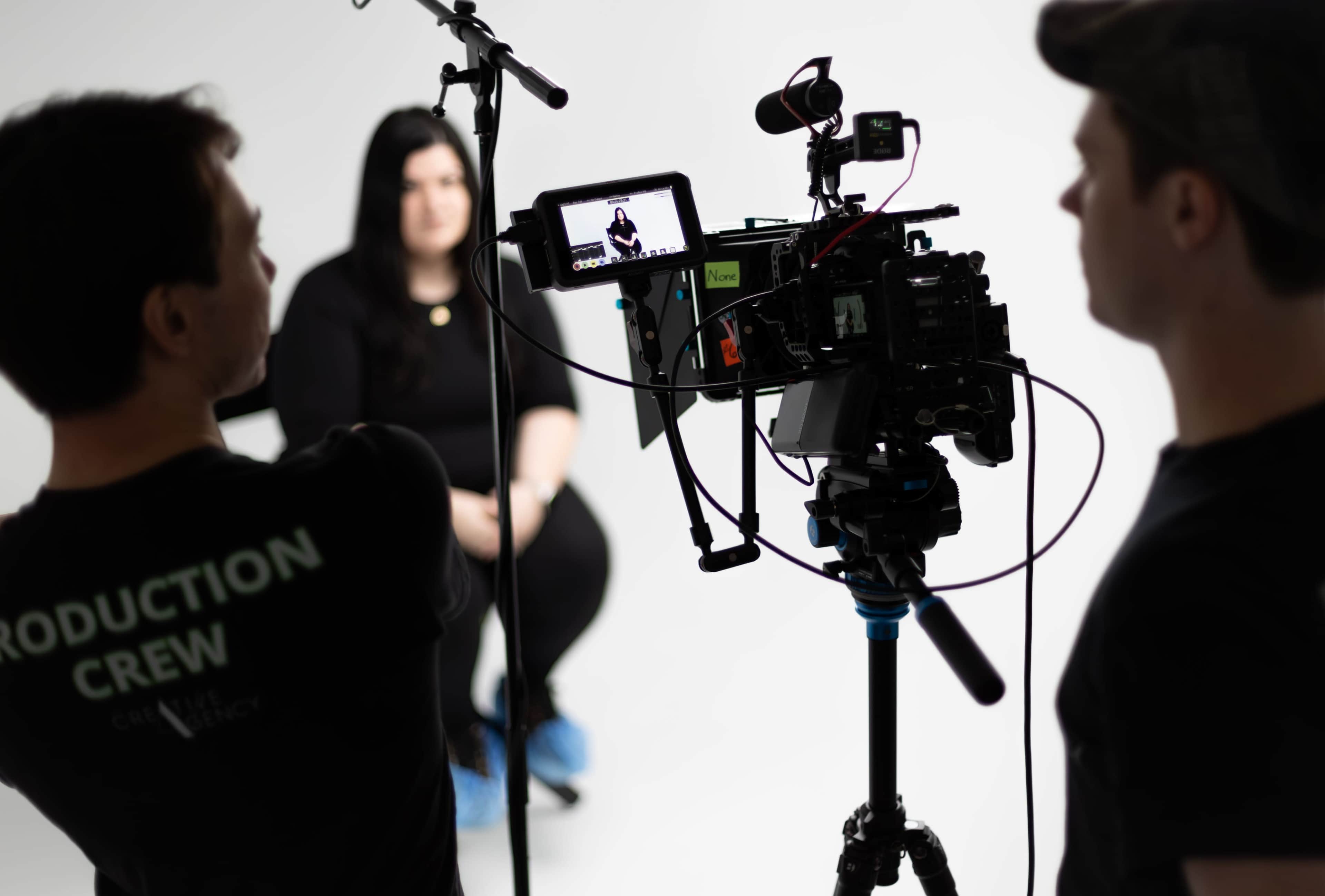 A camera crew is filming an interview with a woman against a plain white backdrop.