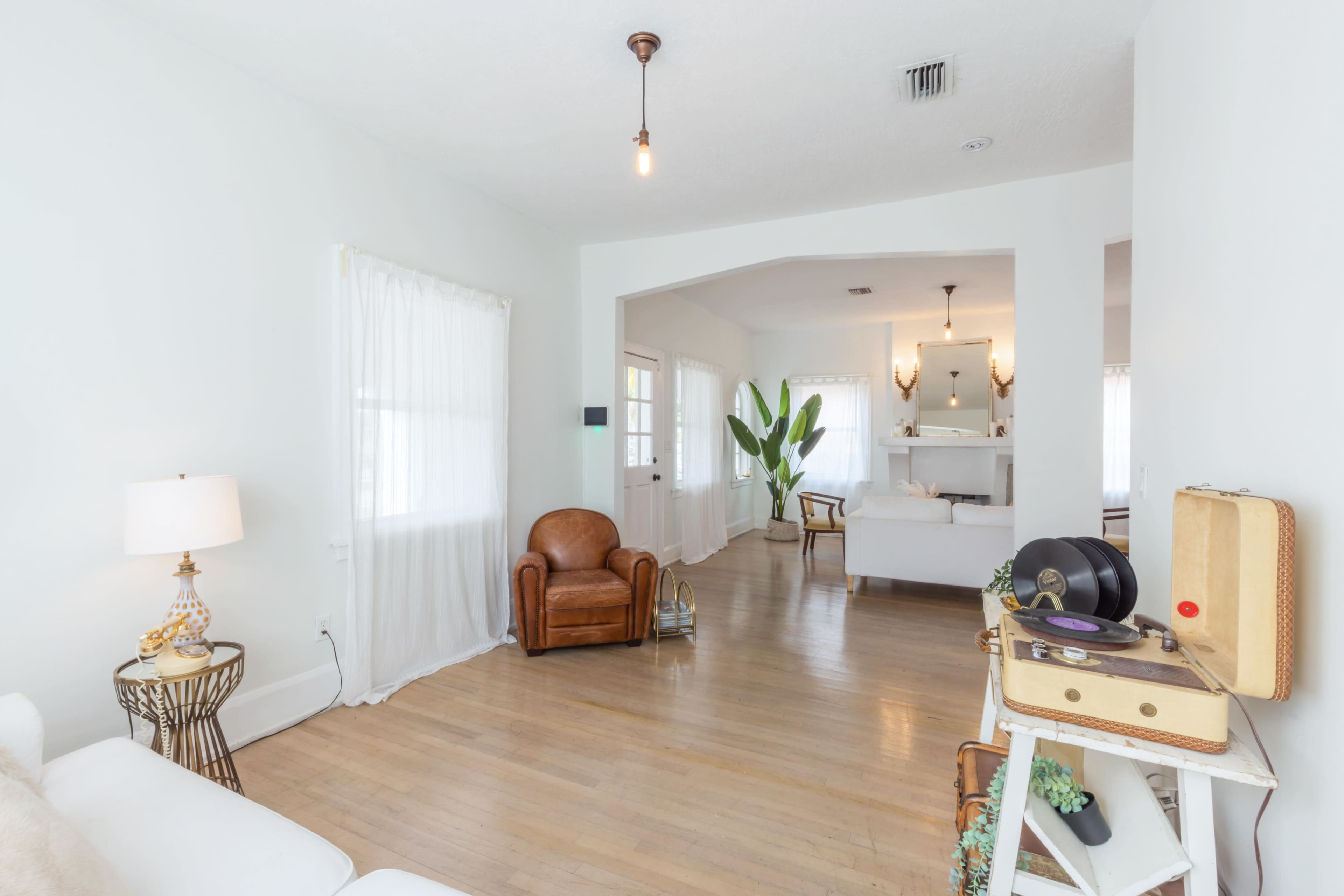 A cozy living room features a brown leather chair, white walls, and light-colored wooden flooring, with a view into a bright adjacent space.
