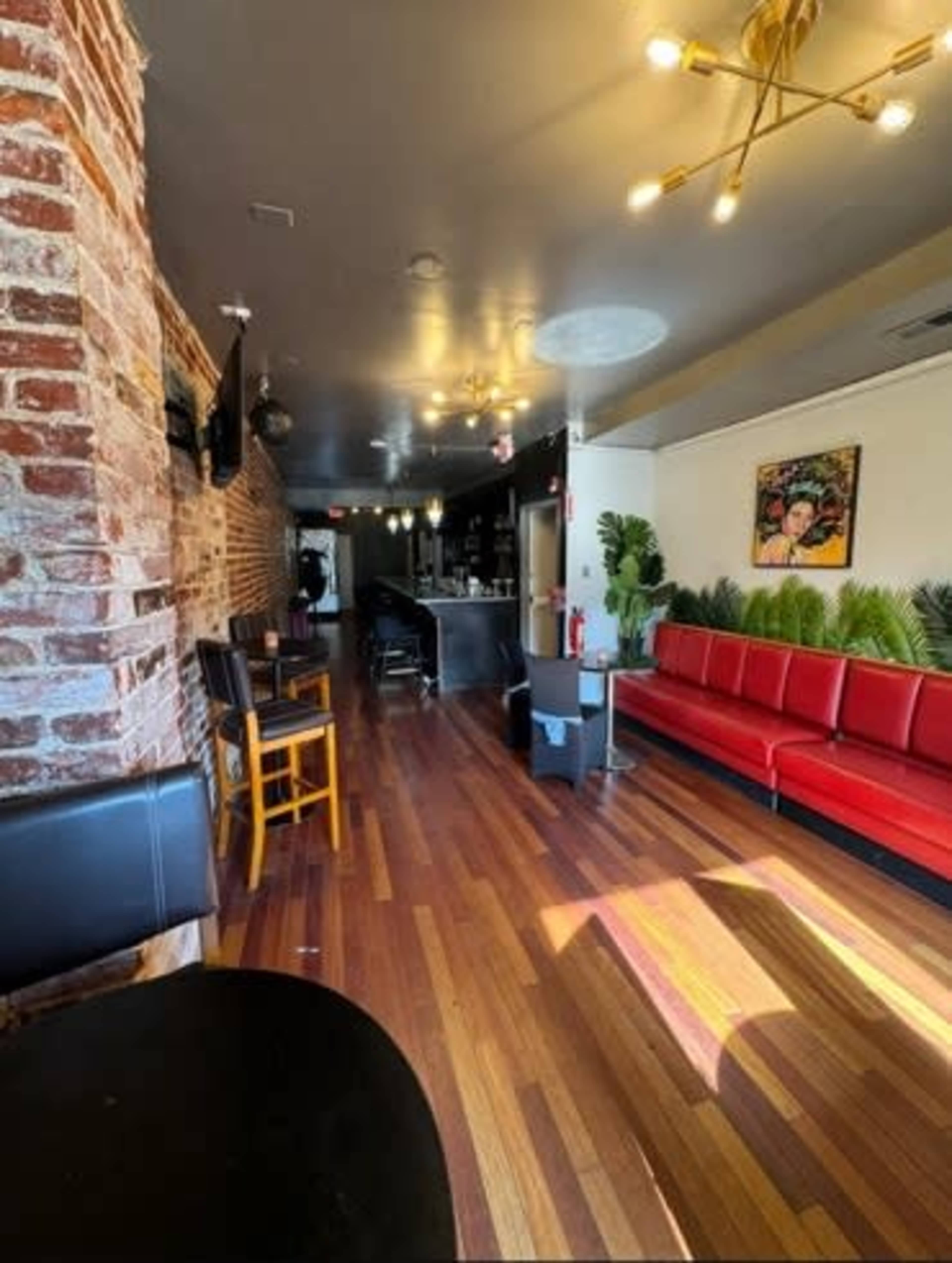 The image shows the interior of a restaurant with a brick wall, wooden flooring, and a long red couch along one side, leading to a bar area in the background.