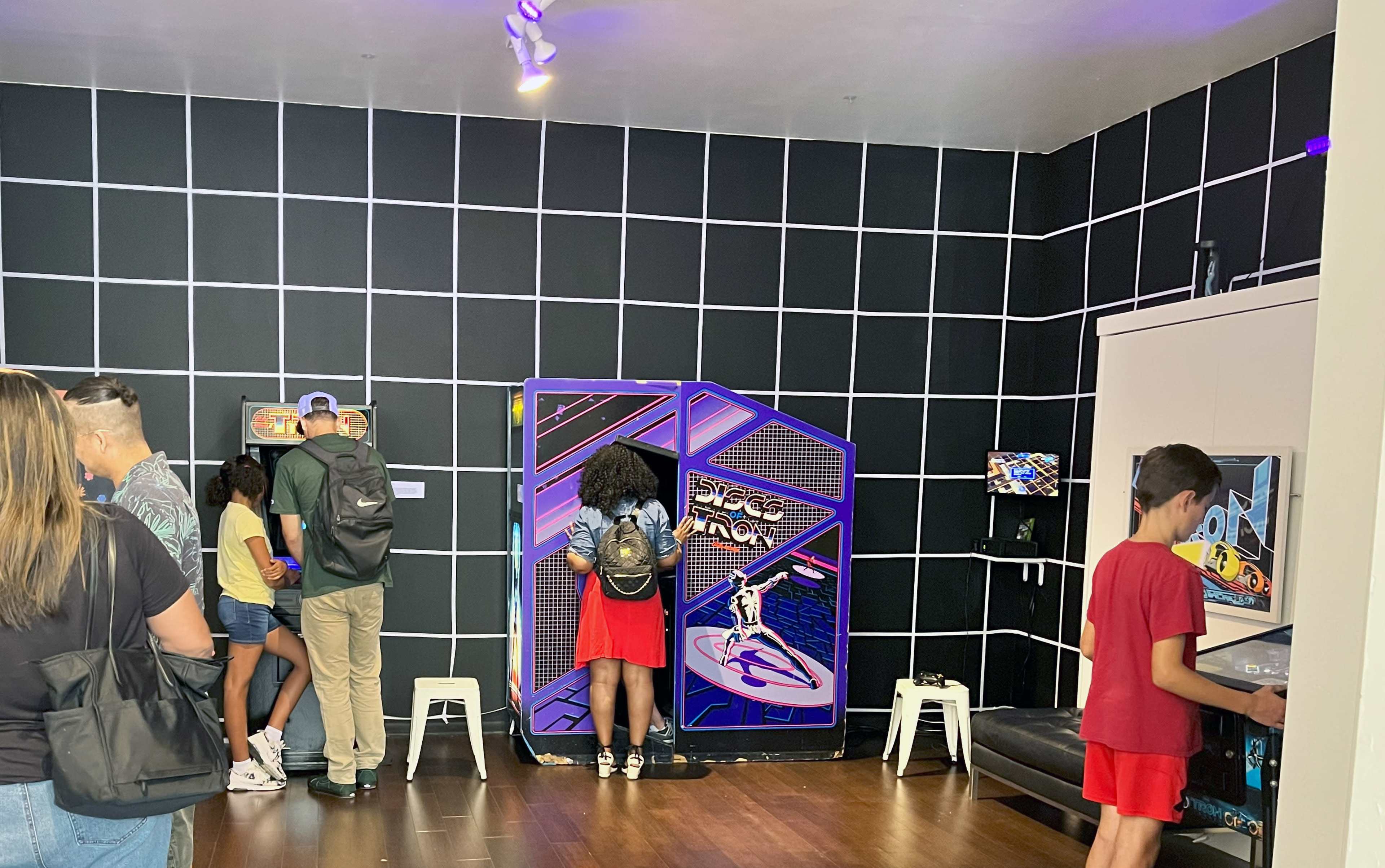 The image shows a group of people interacting with retro arcade games in a room with a grid-patterned black wall.