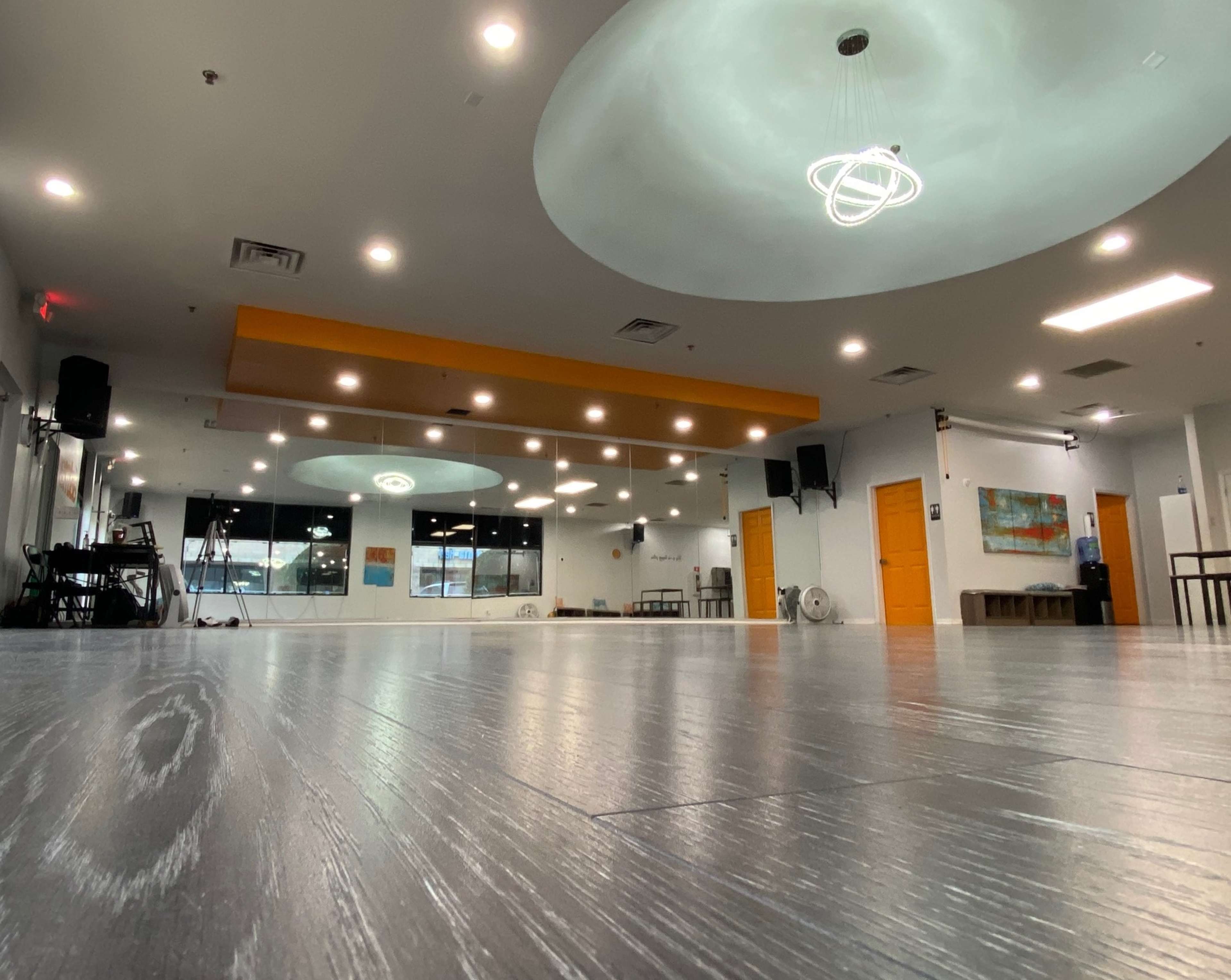 The image shows a spacious event room with a smooth wooden floor, modern lighting fixtures, and orange accent walls.