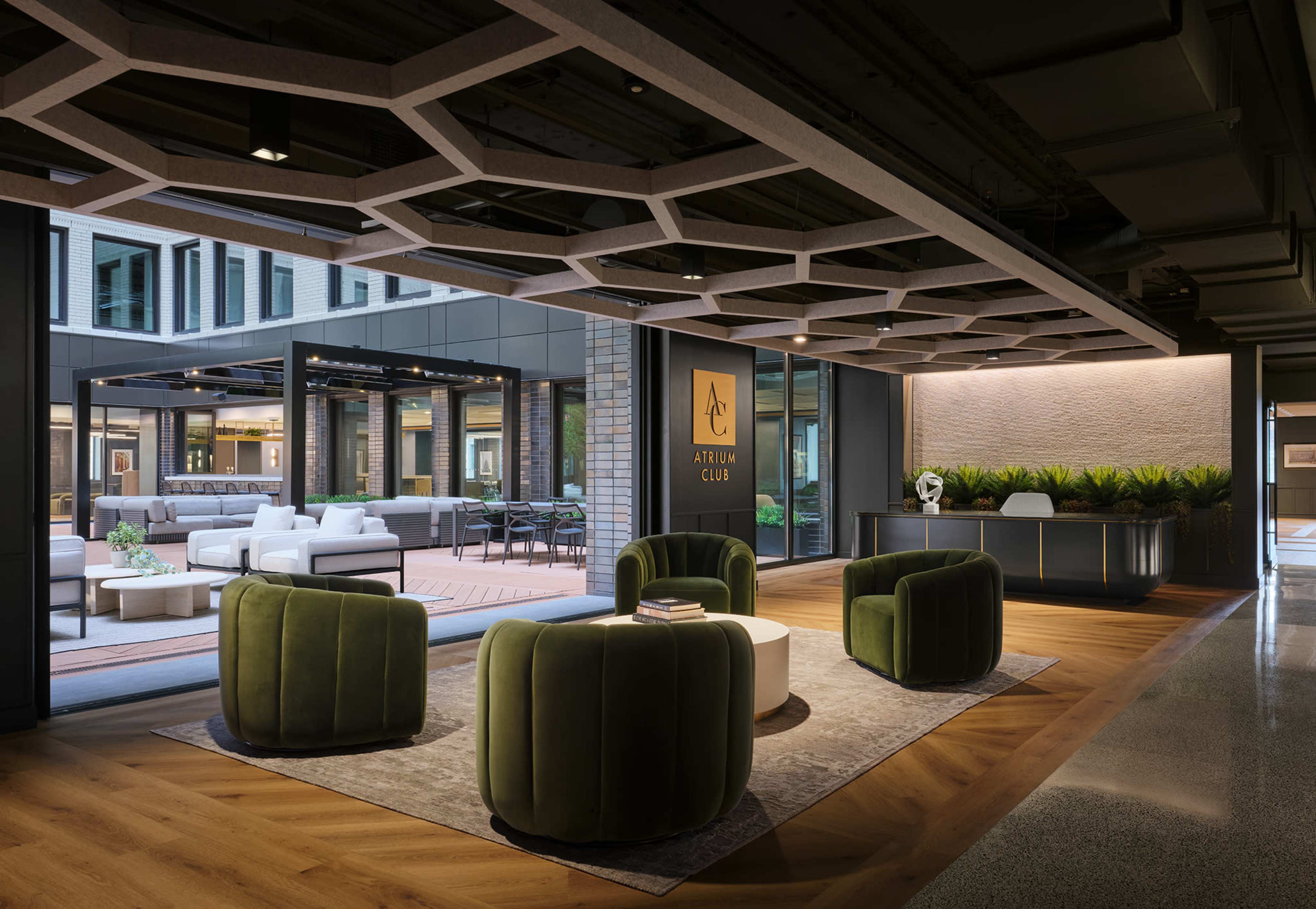 The image shows a modern lobby area with green velvet chairs and a reception desk, overlooking a patio with outdoor seating.