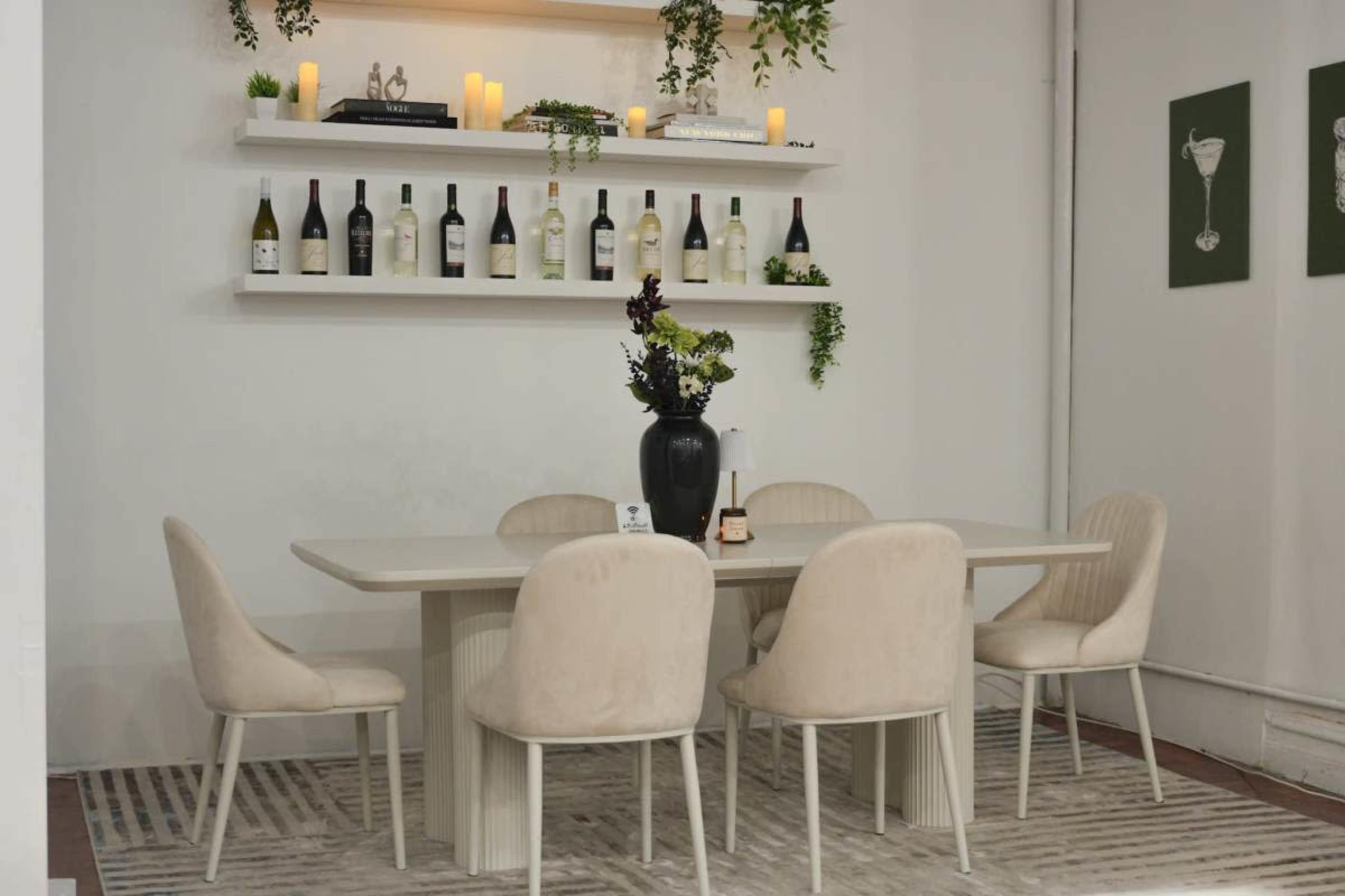 A modern dining area features a light-colored table surrounded by plush chairs, flanked by shelves displaying wine bottles and decorative items.