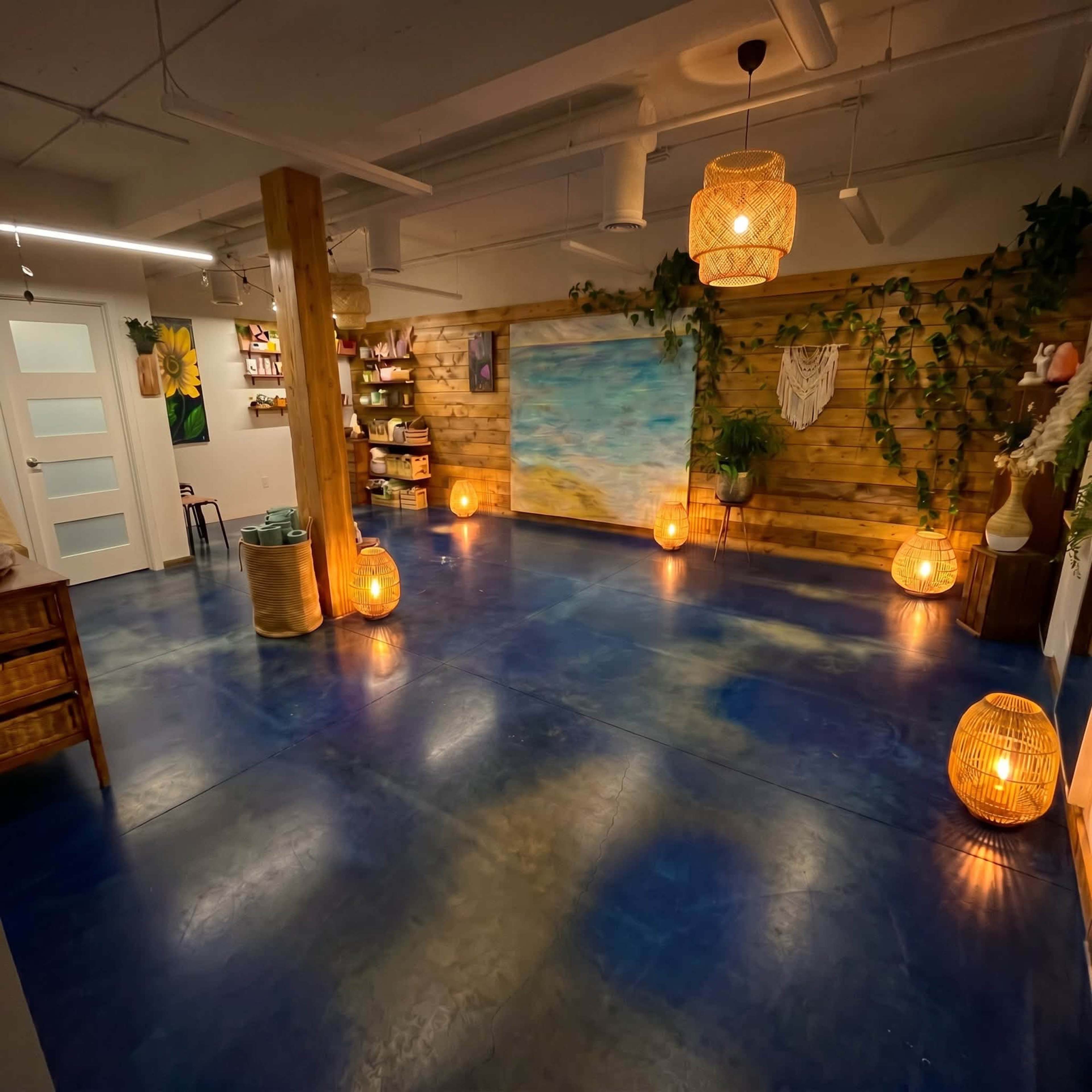 The image shows a spacious room with a blue floor, wooden walls, decorative plants, and warm lanterns providing ambient lighting, along with a large painting of a seascape.
