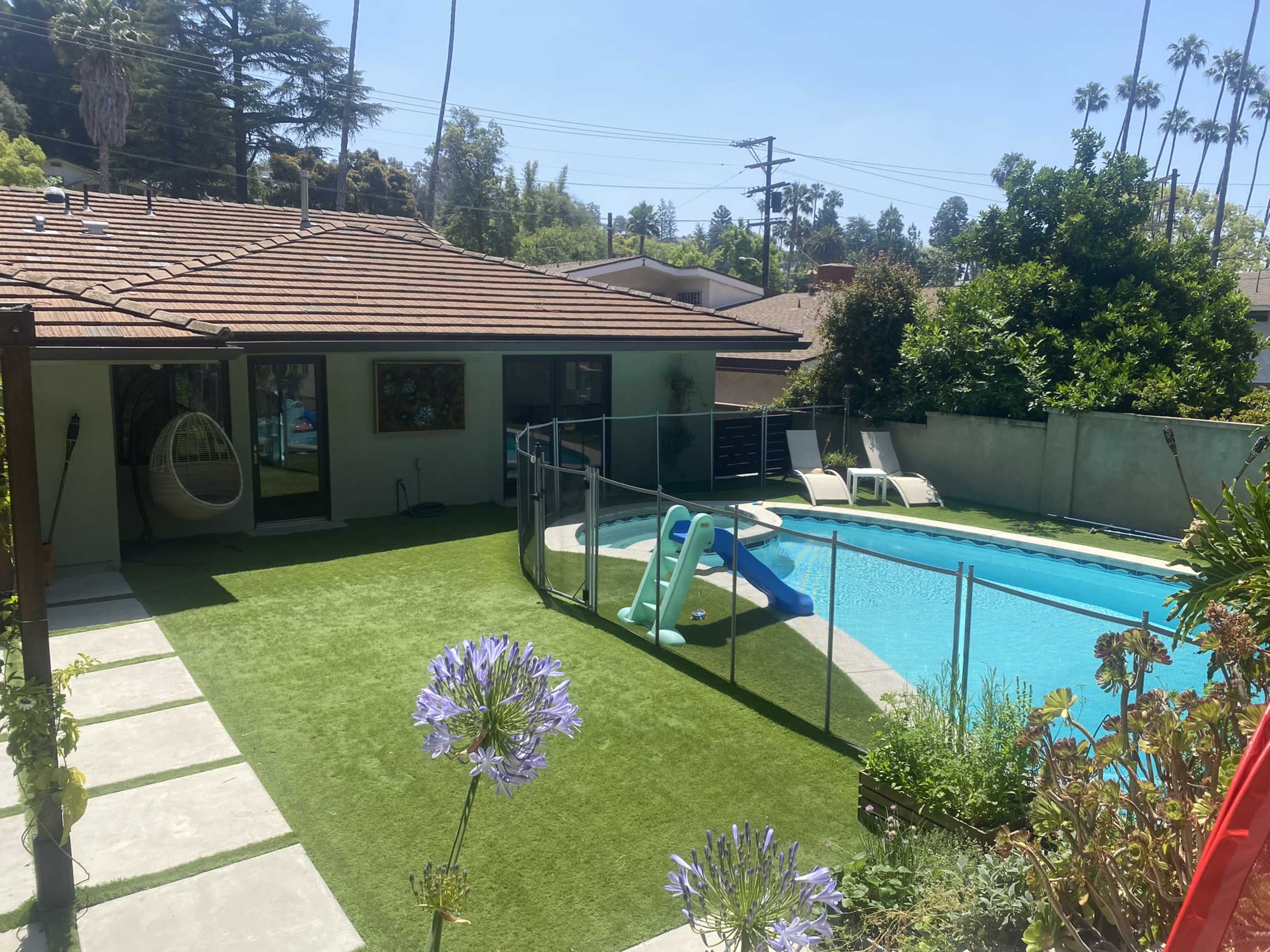 A sunny backyard features a swimming pool with a slide, surrounded by a patio with lounge chairs and a grassy area, set against a backdrop of palm trees.