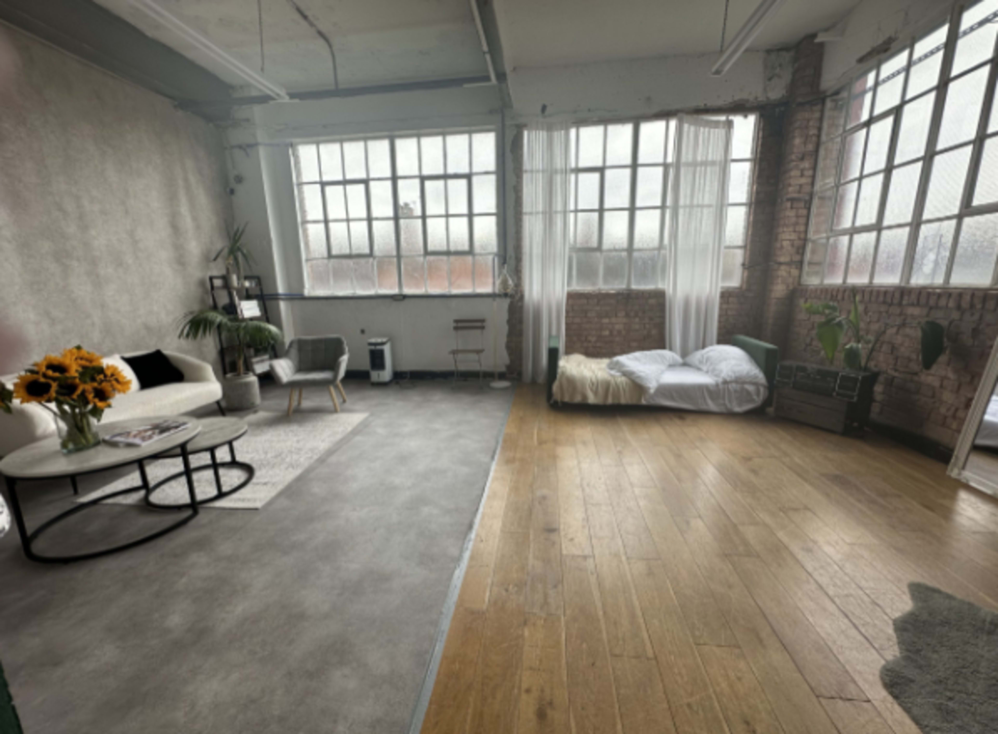 The image shows a spacious, industrial-style room with large windows, a seating area with a white couch, and a bed set up on the floor.