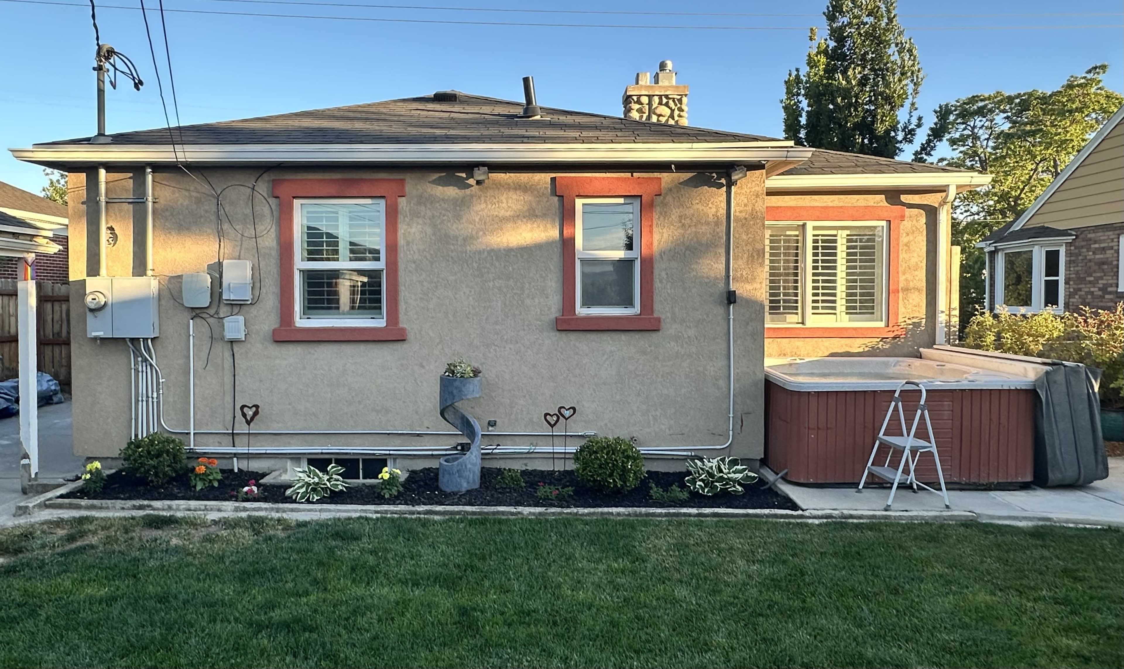 The image shows the back of a house with a hot tub, landscaped garden, and decorative features near the windows.