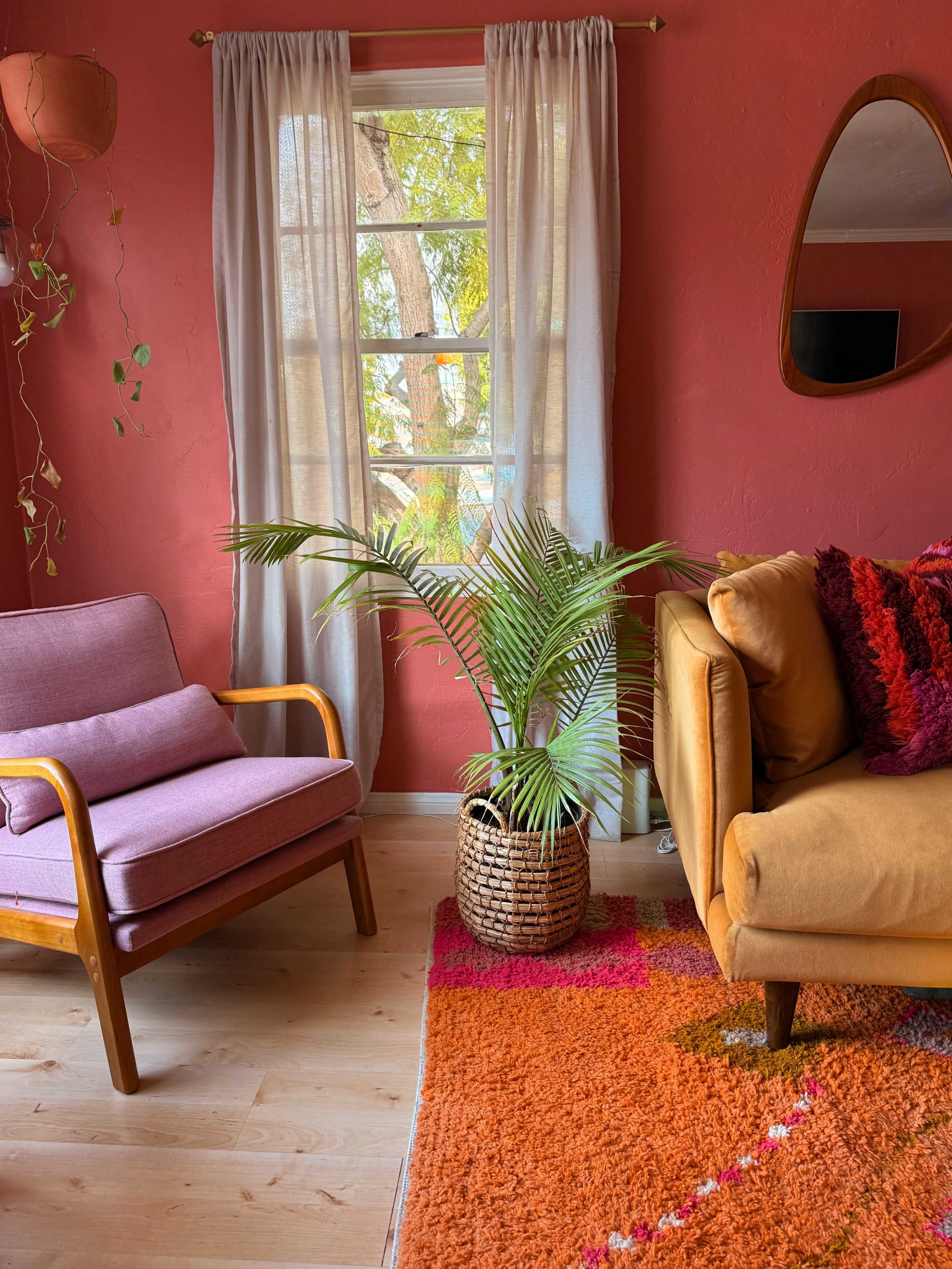 The image depicts a cozy living room with a pink armchair, a mustard-colored sofa, a large woven plant pot with a palm, and a colorful area rug, all set against a vibrant red wall.