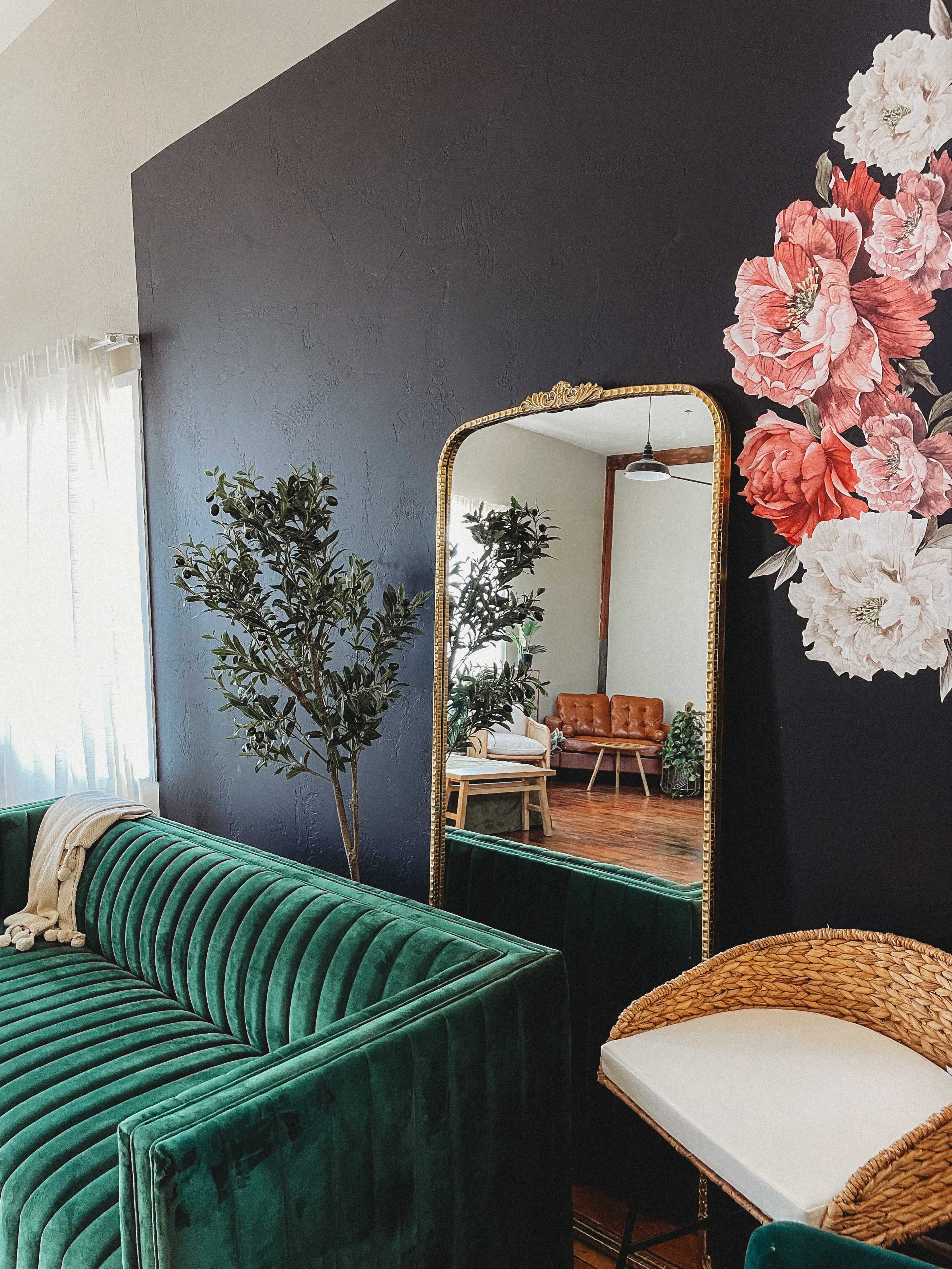 A cozy living room featuring a green velvet sofa, a large mirror with an ornate frame, and floral wall art alongside potted plants.