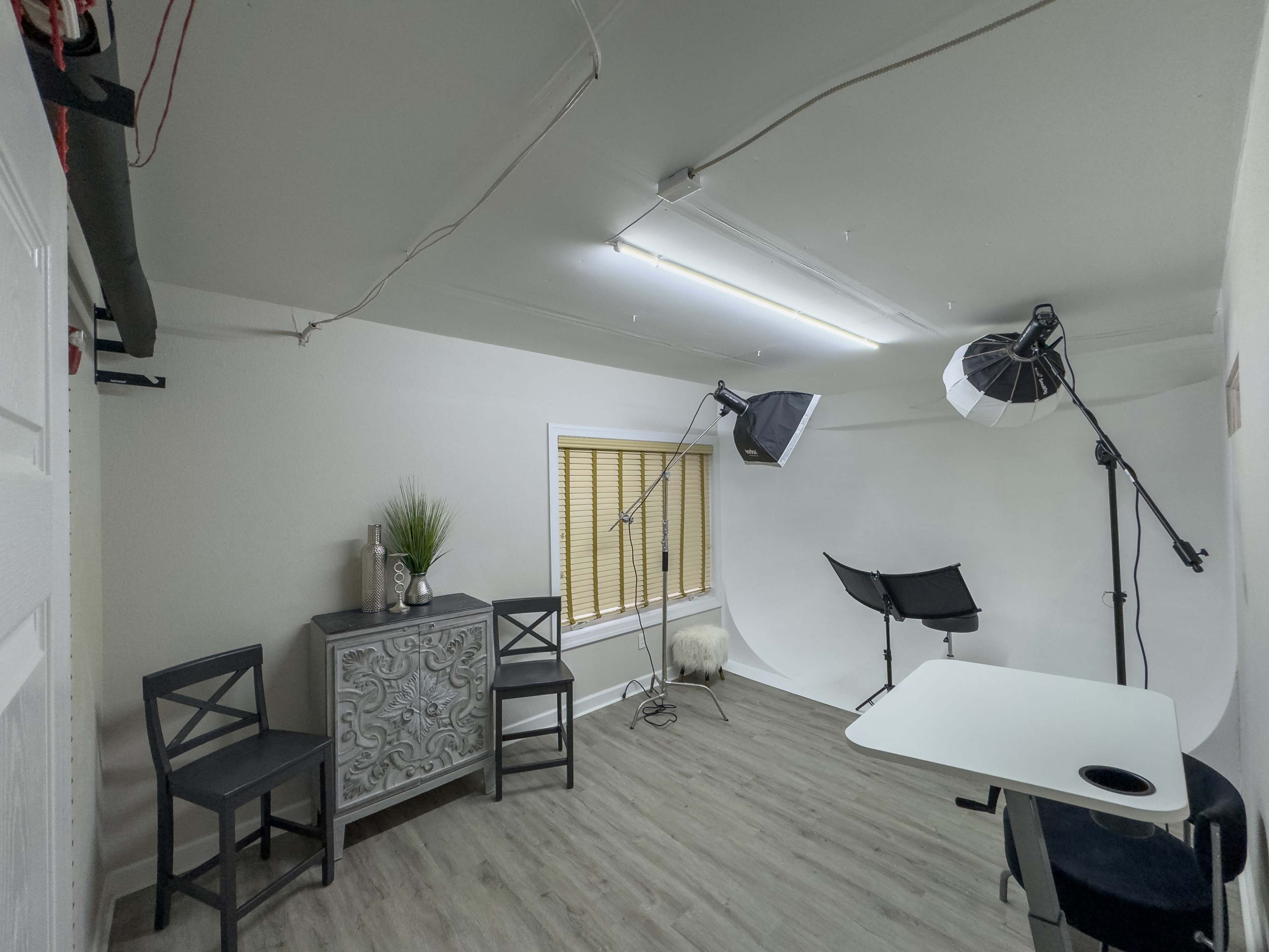 A small, well-lit studio space featuring a table, two chairs, a decorative cabinet, and photography equipment.