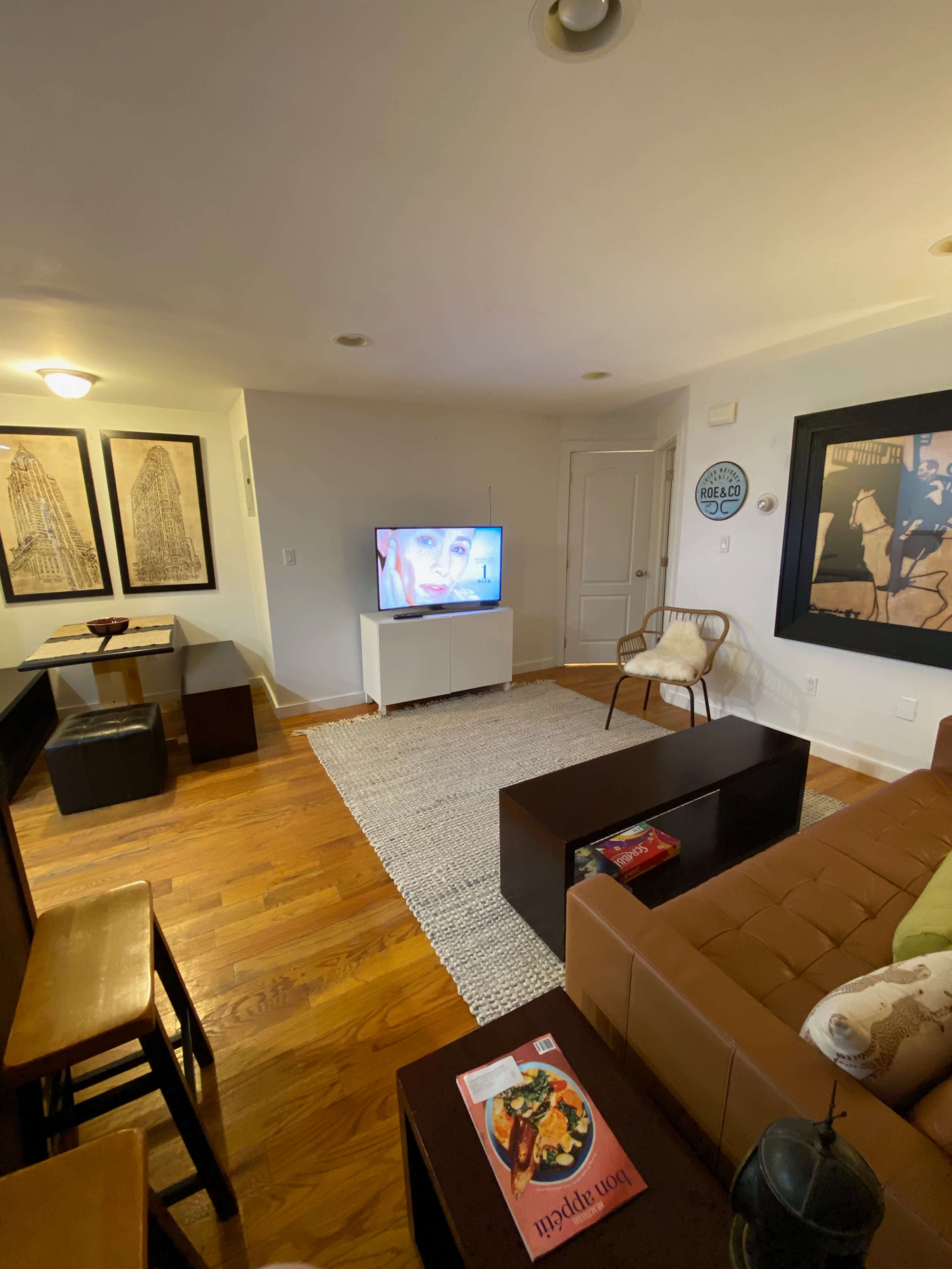 A living room with a television on a white cabinet, a brown sofa, a large rug, and wooden chairs arranged around a coffee table.