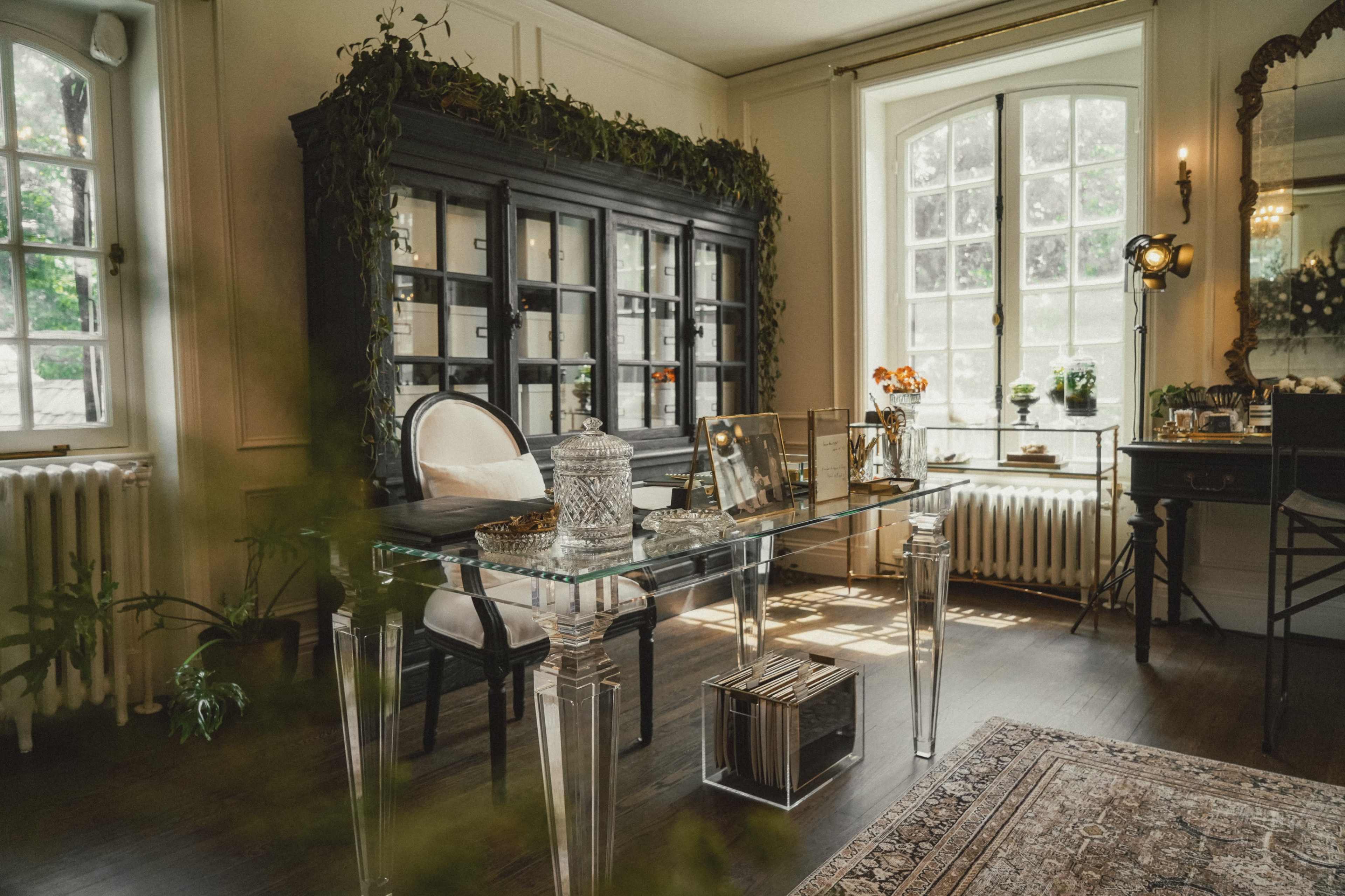 The image shows a stylish office space featuring a glass desk, a black cabinet with shelves, and large windows allowing natural light.