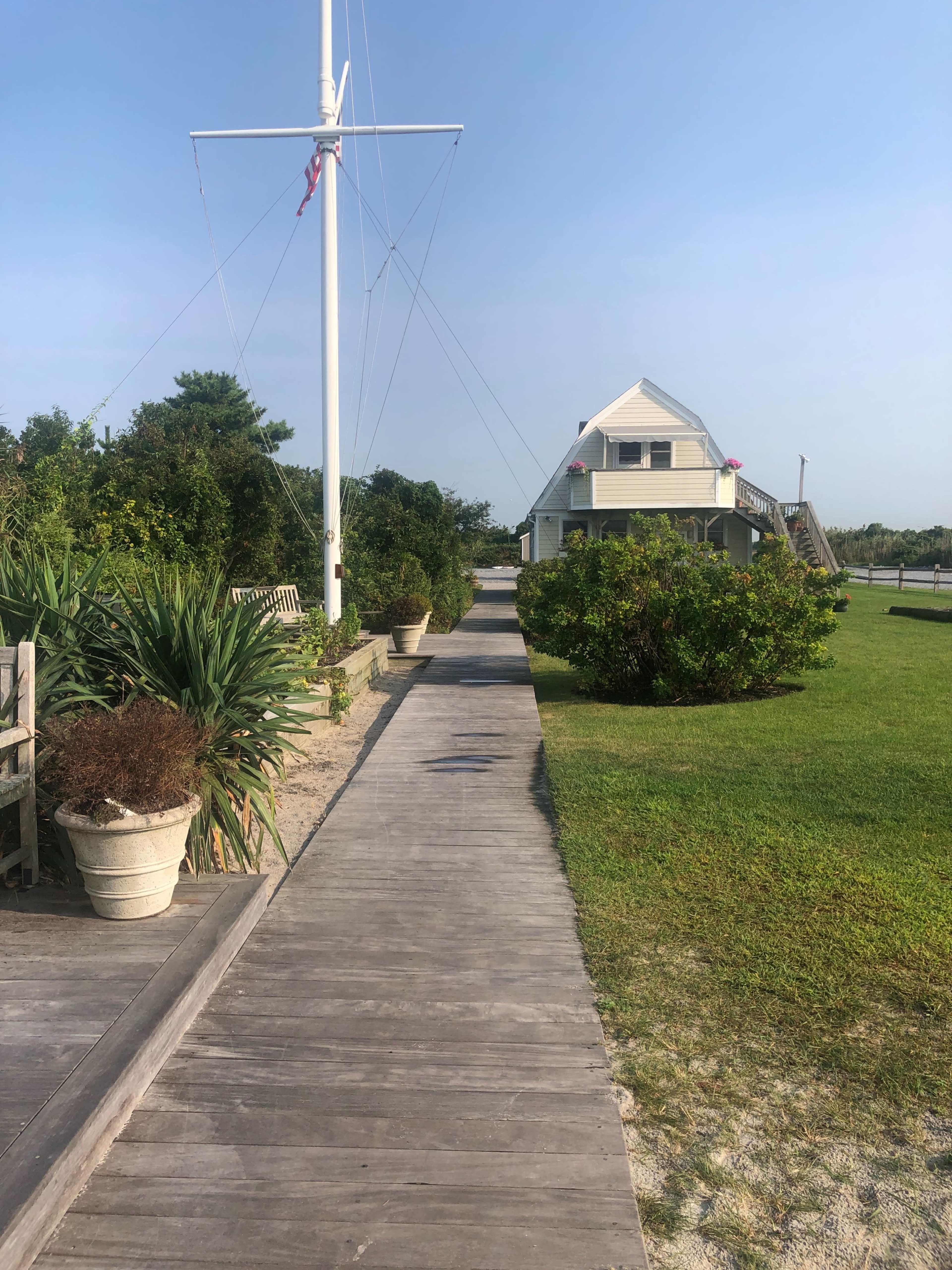 A wooden pathway lined with potted plants leads to a two-story house near the shore.