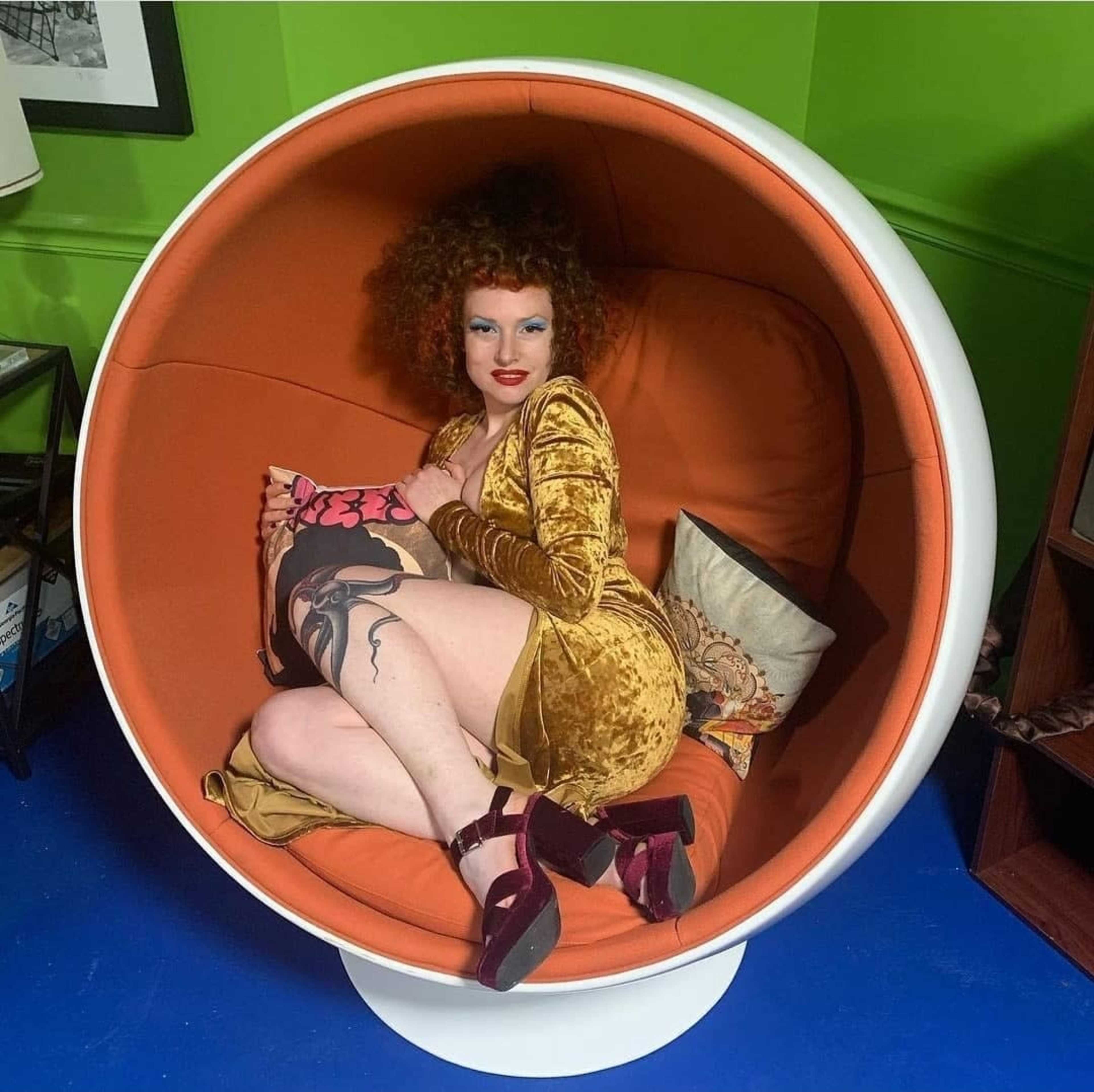 A person with curly hair sits inside a round, orange chair, holding a decorative pillow and wearing a yellow dress.