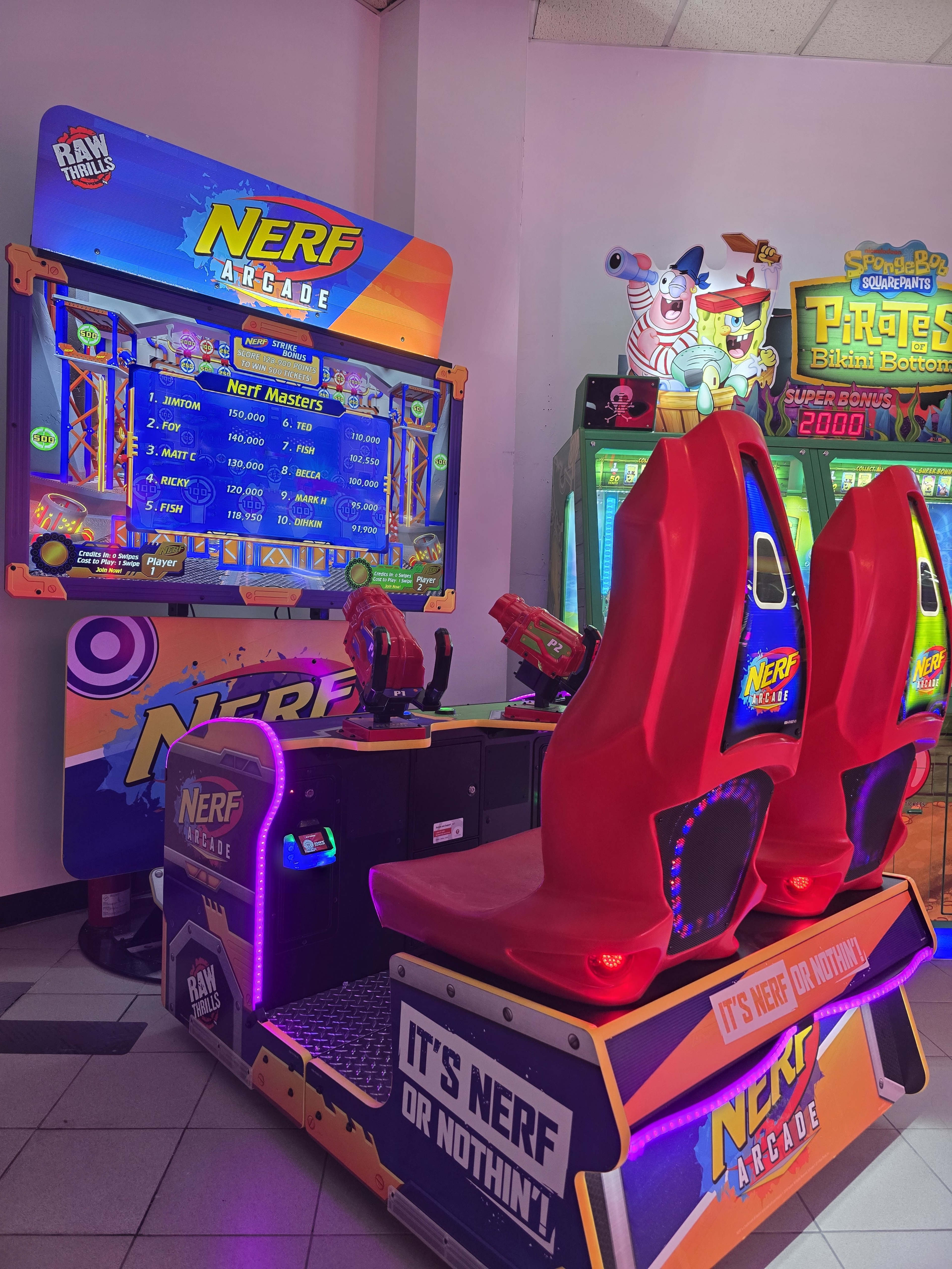 The image shows a brightly lit Nerf arcade game setup featuring two red seating stations and a vibrant screen displaying the game options.