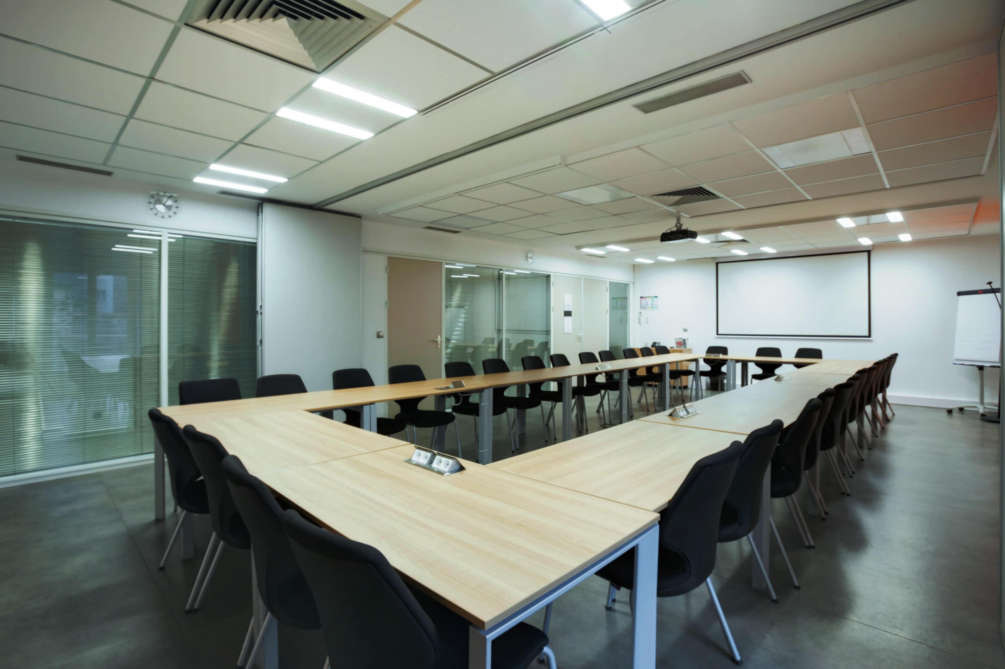 A large conference room features a U-shaped table surrounded by black chairs, with a projector screen at one end and glass walls.