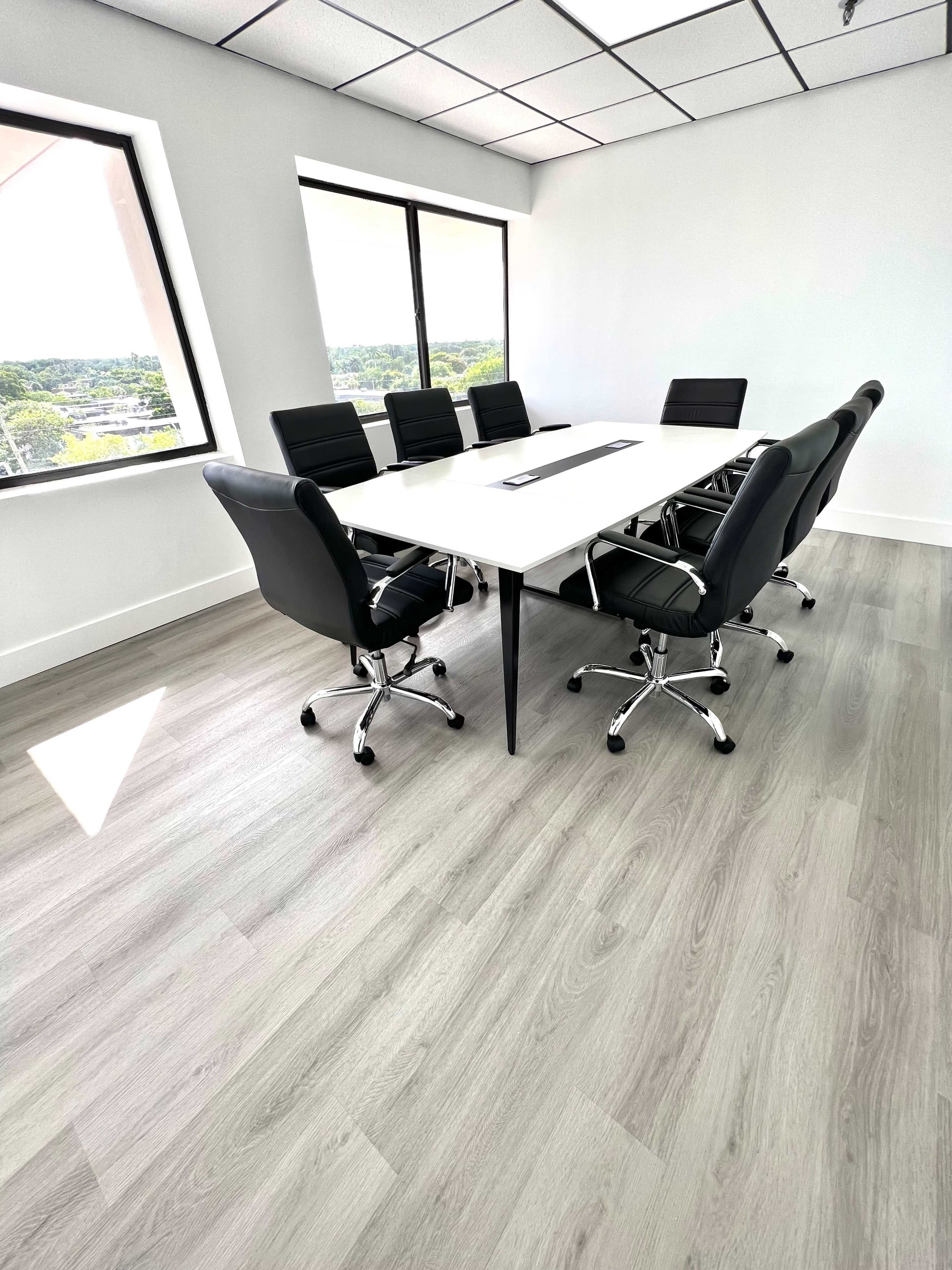 A modern conference room features a large white table surrounded by black ergonomic chairs and large windows with a view.