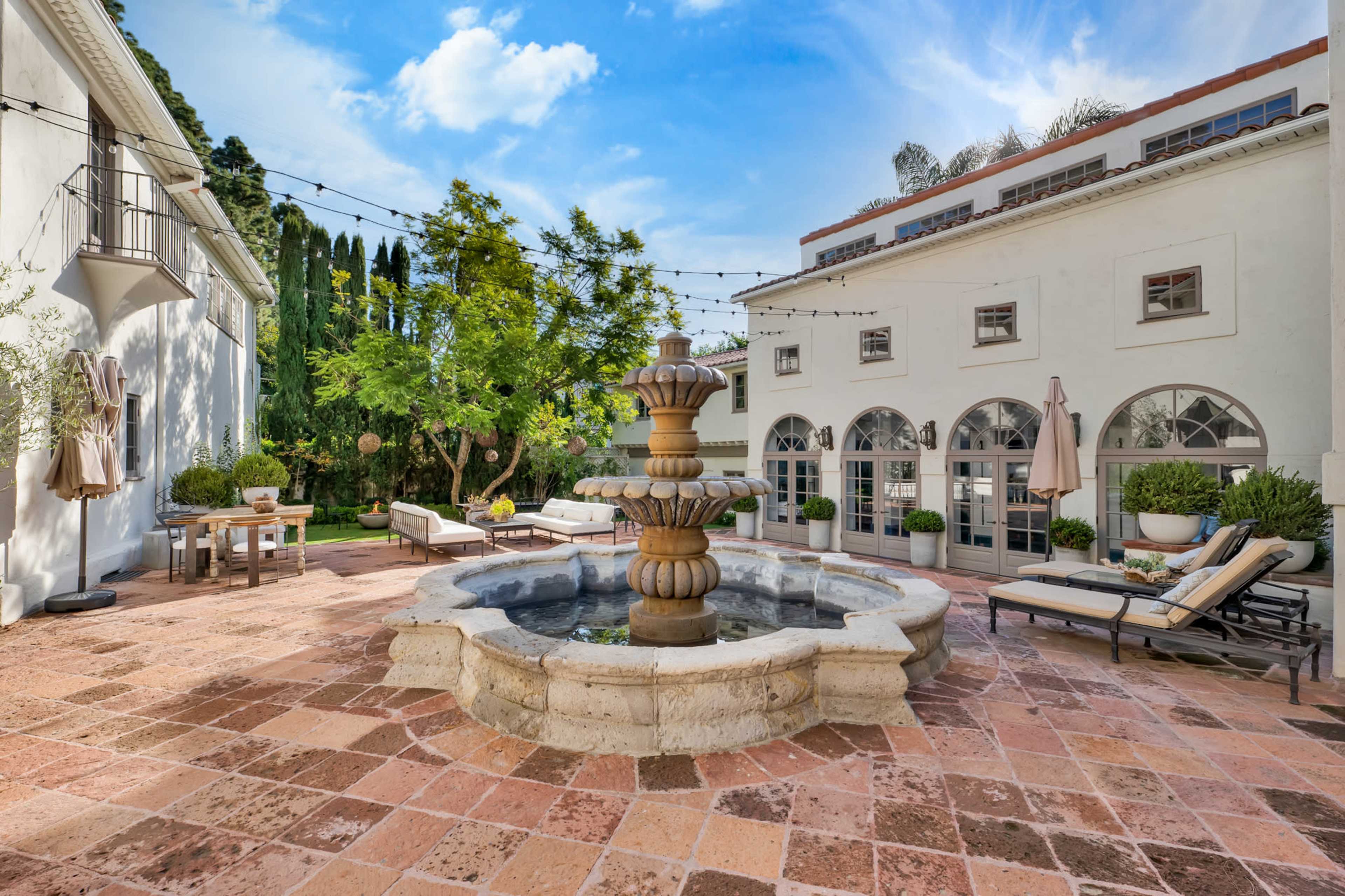A stone fountain creates a central feature in a brick patio surrounded by lounge chairs and greenery.