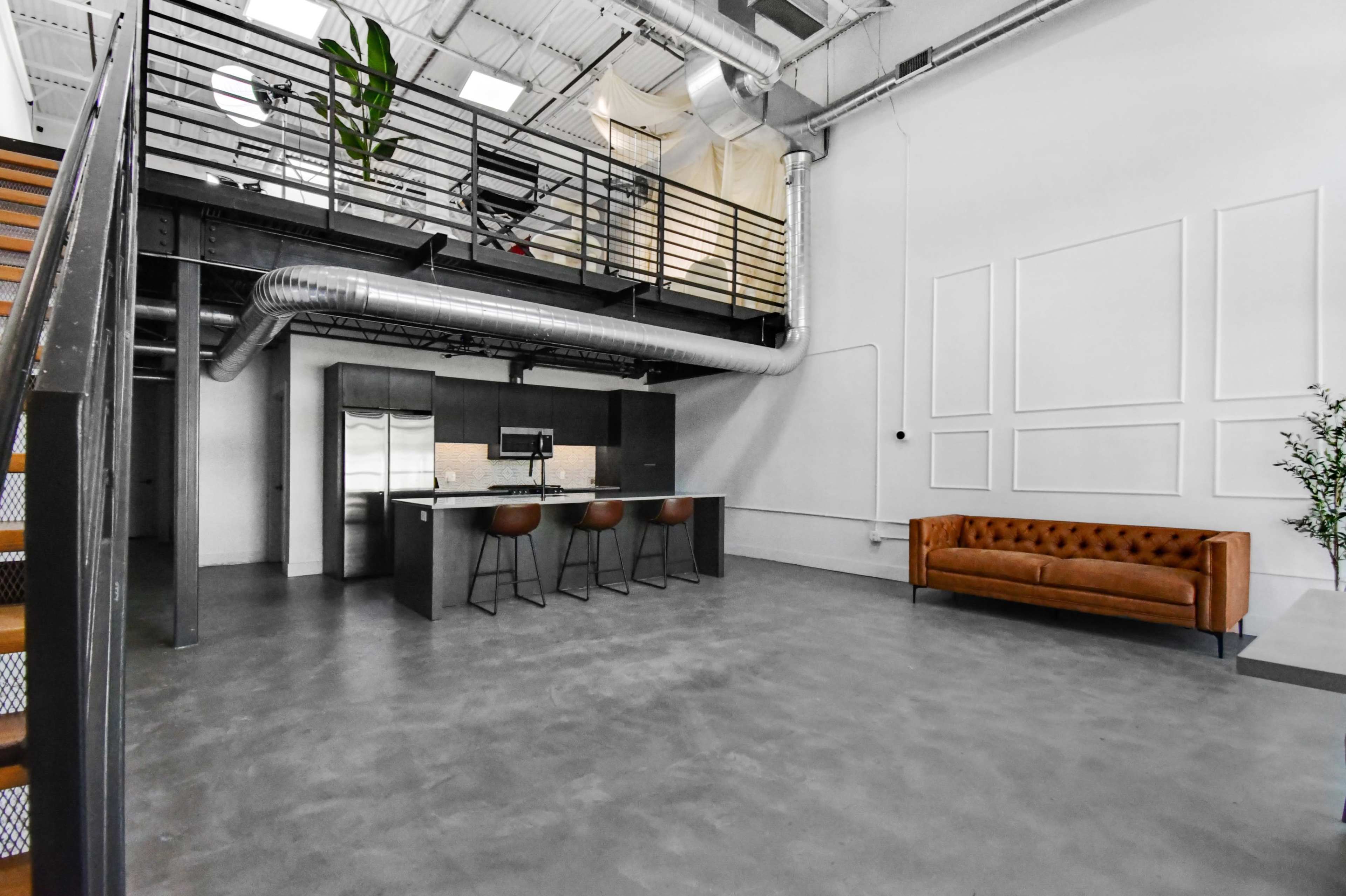 The image shows an industrial-style interior with a kitchen island featuring bar stools, a leather sofa, and a staircase leading to an upper level.