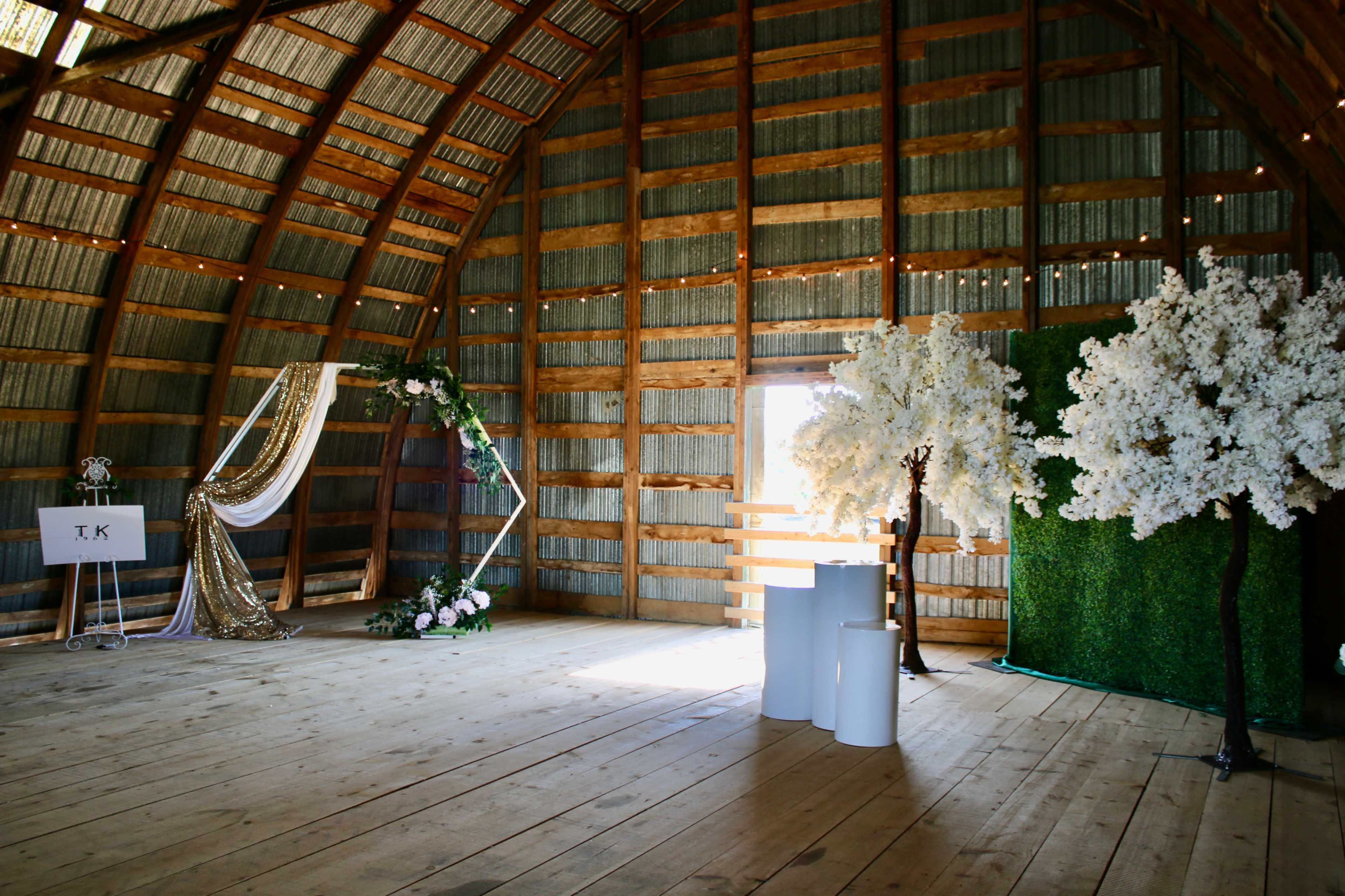 The interior of a rustic barn features decorative floral arrangements, a fabric drapery display, and a backdrop of artificial trees.