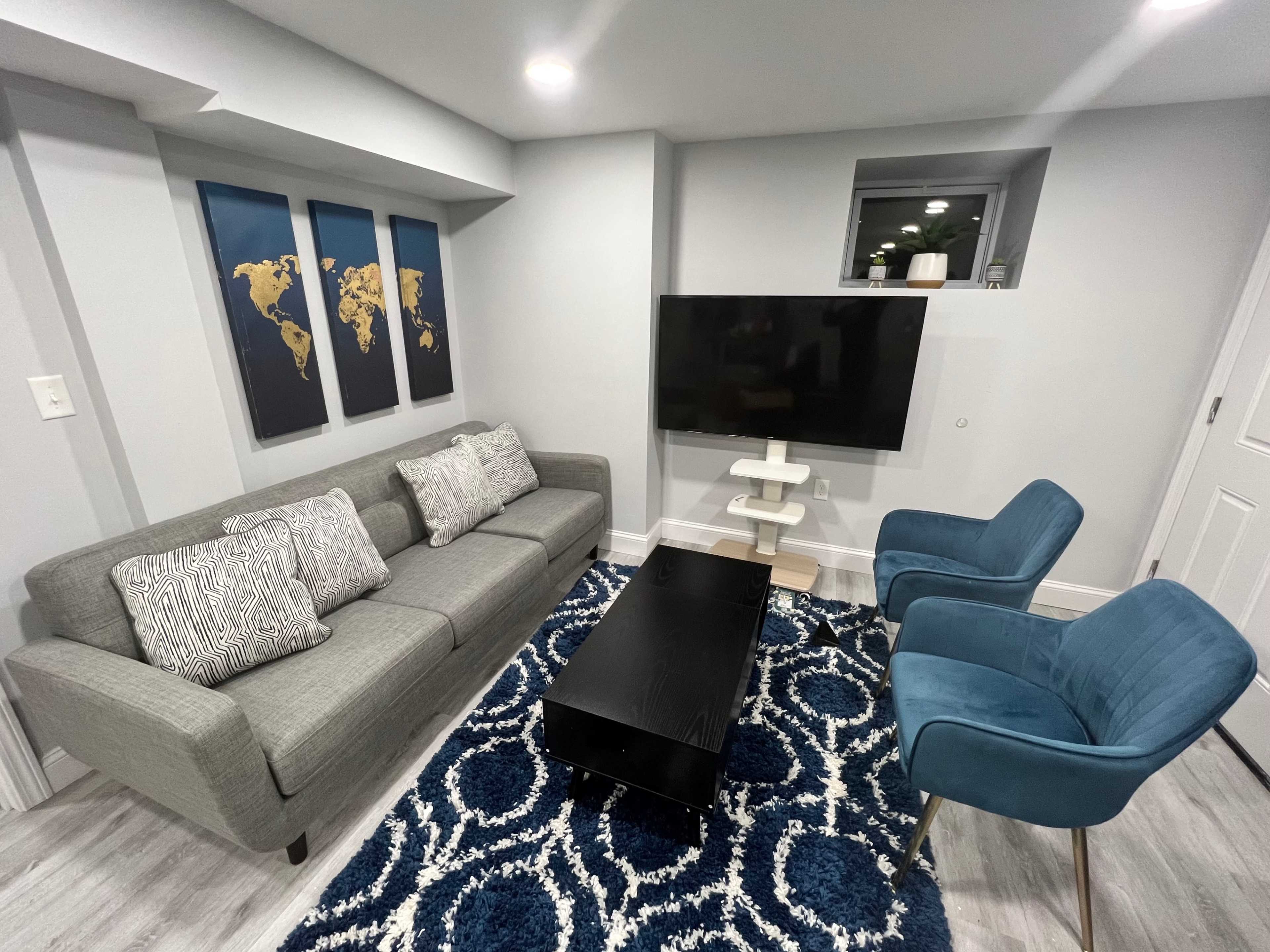 A modern living room featuring a gray sofa with decorative pillows, a black coffee table, two blue chairs, a TV mounted on the wall, and a world map art piece.