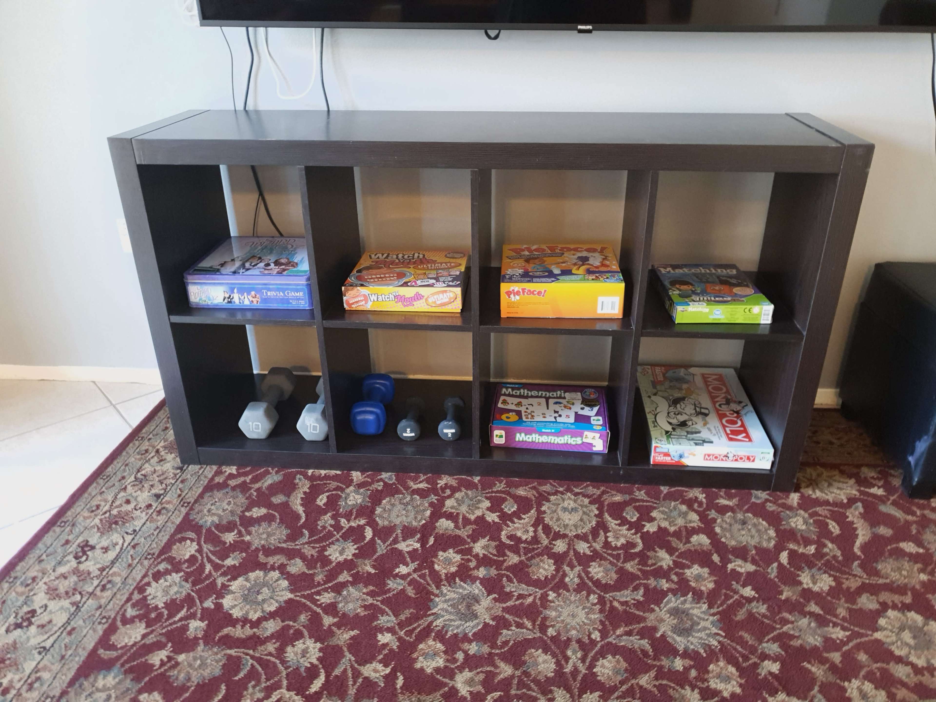 A rectangular shelf unit displays various board games and a set of dumbbells in a living room setting.