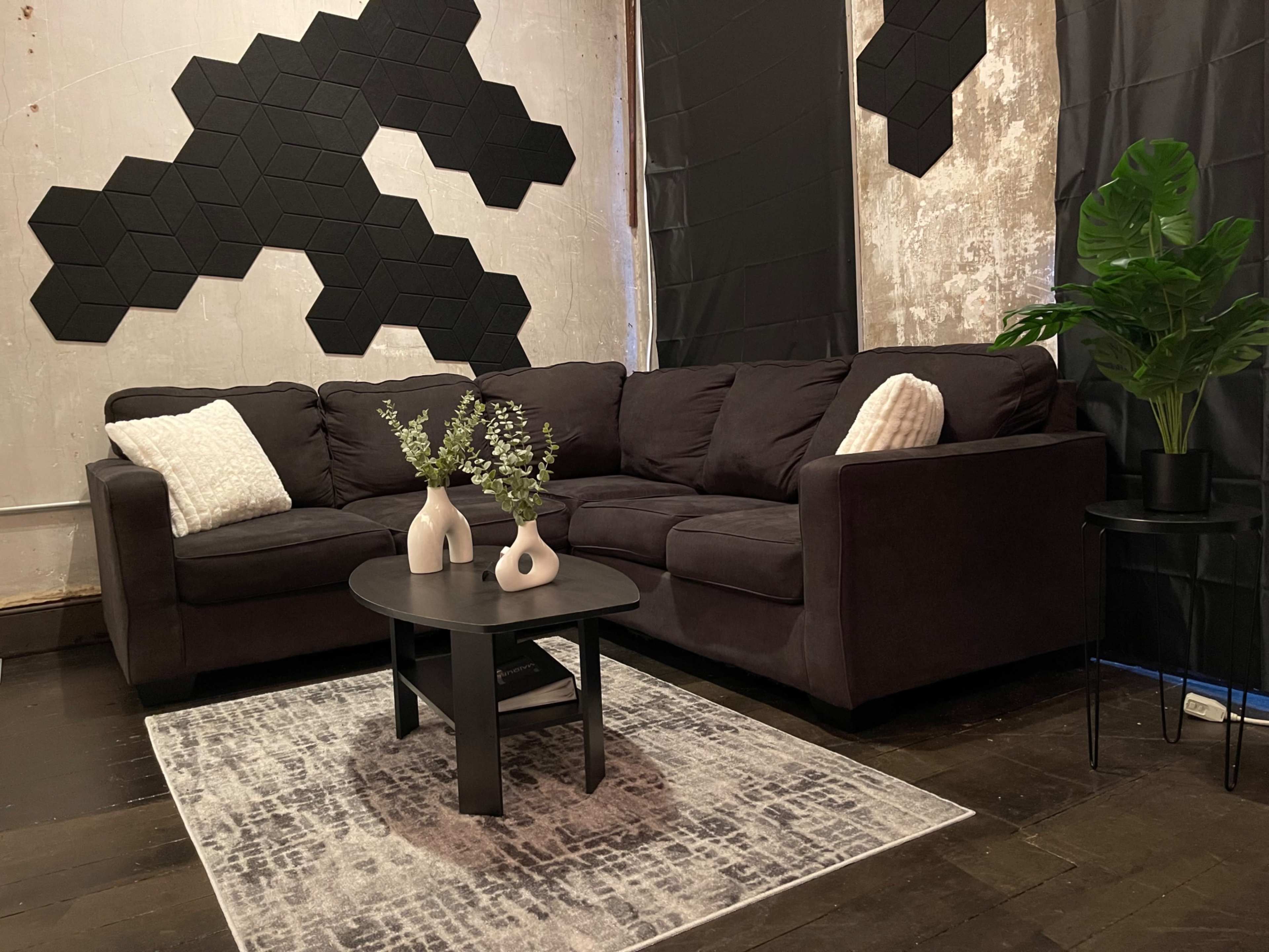 A dark sectional sofa with a round black coffee table and vases of greenery sits on a patterned rug in a room with textured walls and hexagonal wall art.