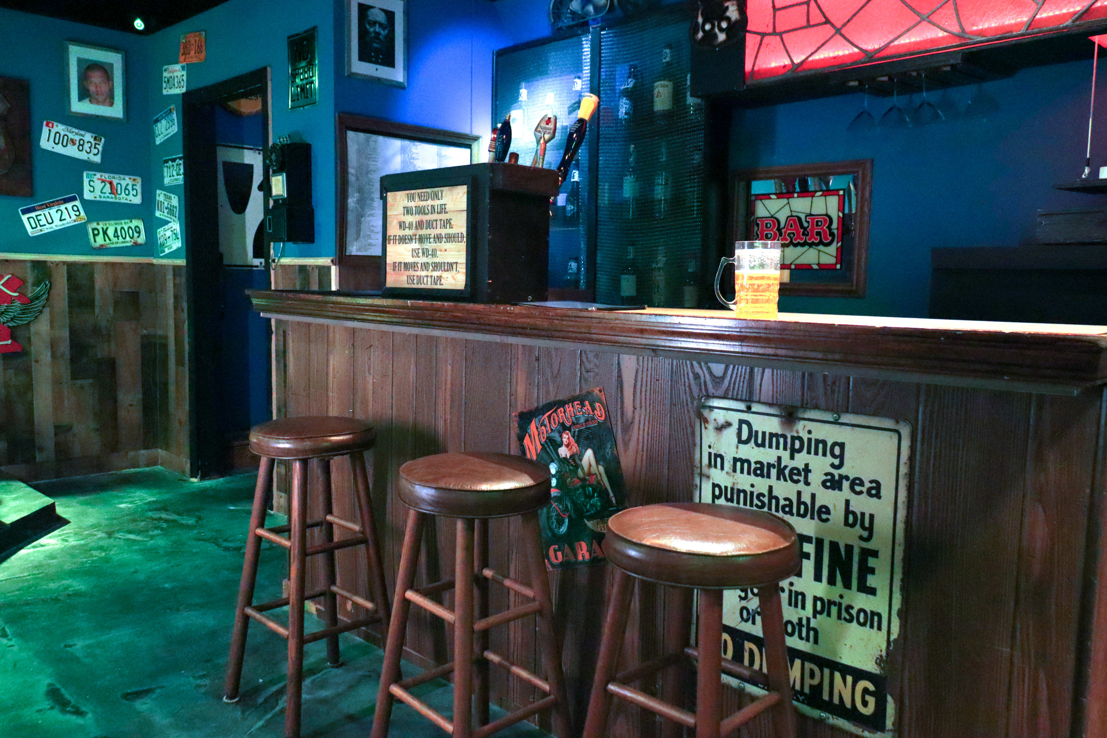 The image shows a wooden bar with three empty stools, neon decorations, and various signs on the walls.