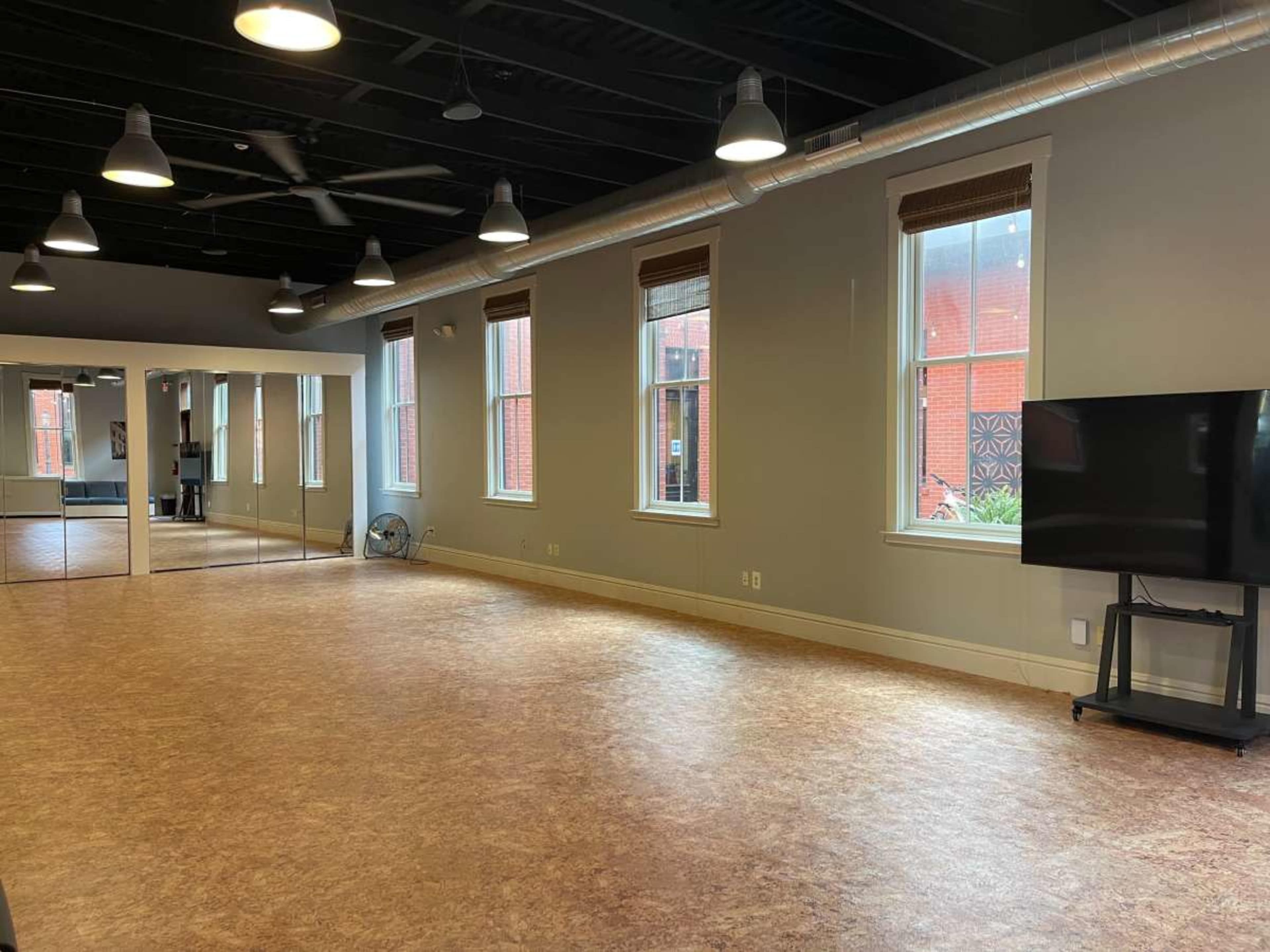 The image shows a spacious room with wooden flooring, large windows, and a mirror along one wall, equipped with a television on a stand.