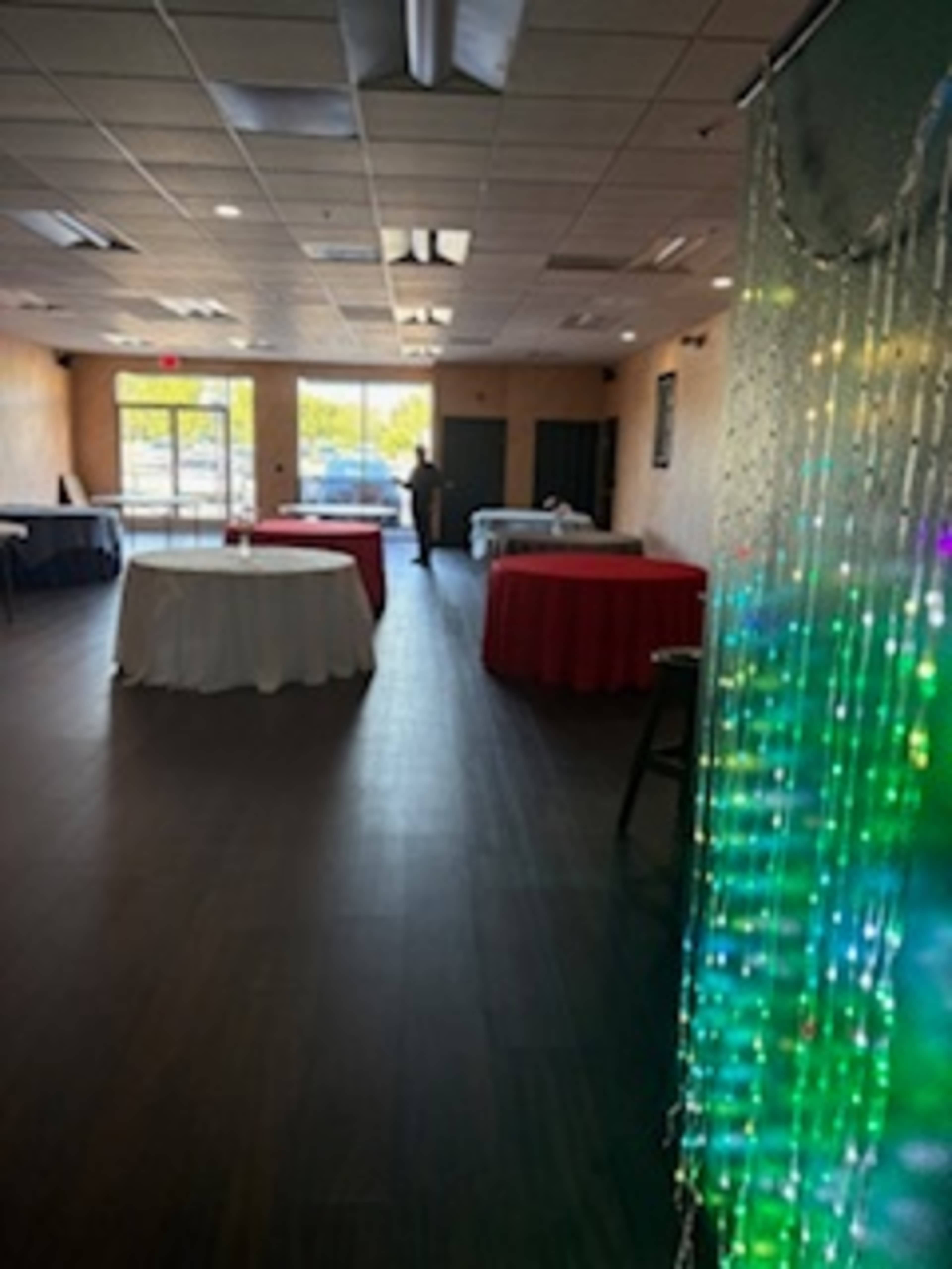The image shows an empty event space with tables covered in white and red tablecloths and a colorful light display on one side.