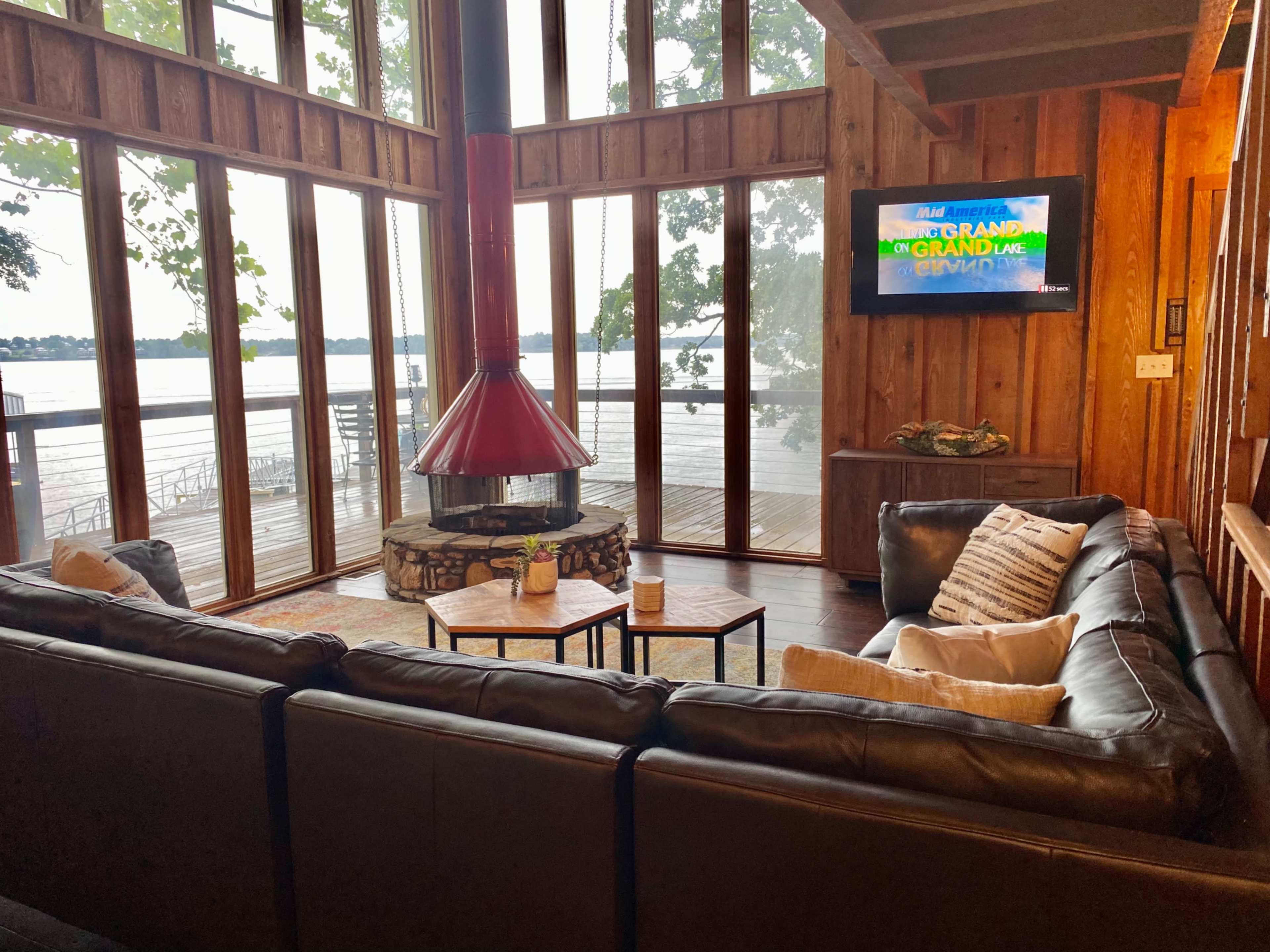 The image shows a cozy living room with a circular fireplace, large windows overlooking a lake, and a sectional sofa arranged around a coffee table.